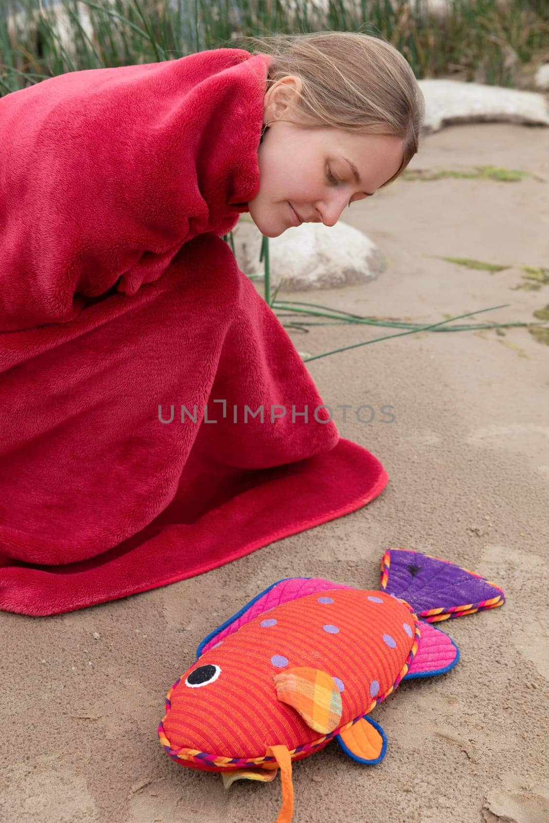 A woman wearing a red blanket leans down to examine a vibrant fish toy resting on sandy ground. The scene captures a tranquil moment by the beach with gentle natural surroundings.