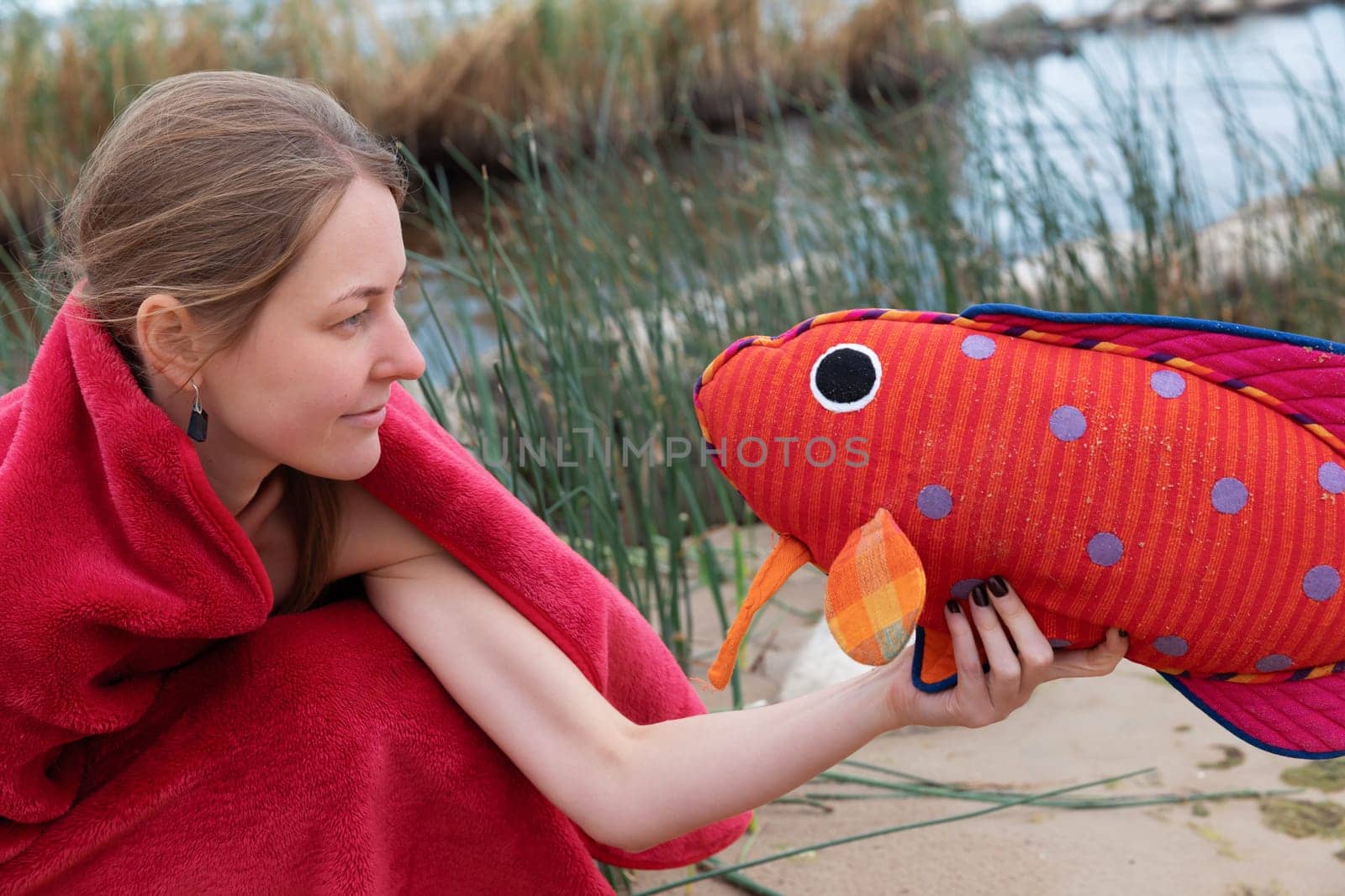 A woman wrapped in a red blanket smiles as she playfully holds a vibrant plush fish at the waters edge. Surrounding grasses and serene water create a peaceful atmosphere.