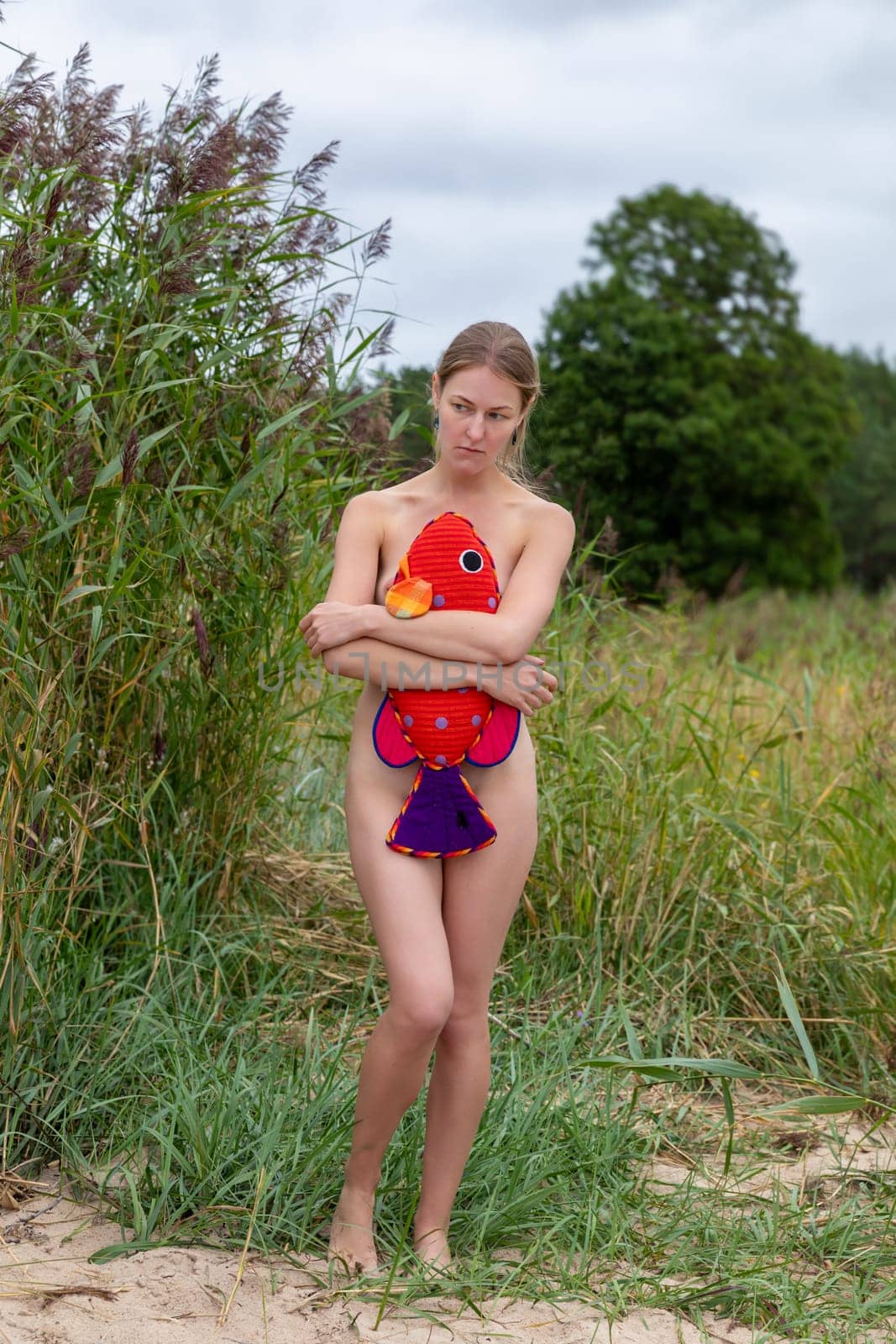 A woman poses by a riverbank wrapped in grass, holding a vibrant plush fish. The atmosphere is serene, with overcast skies and lush greenery surrounding her.