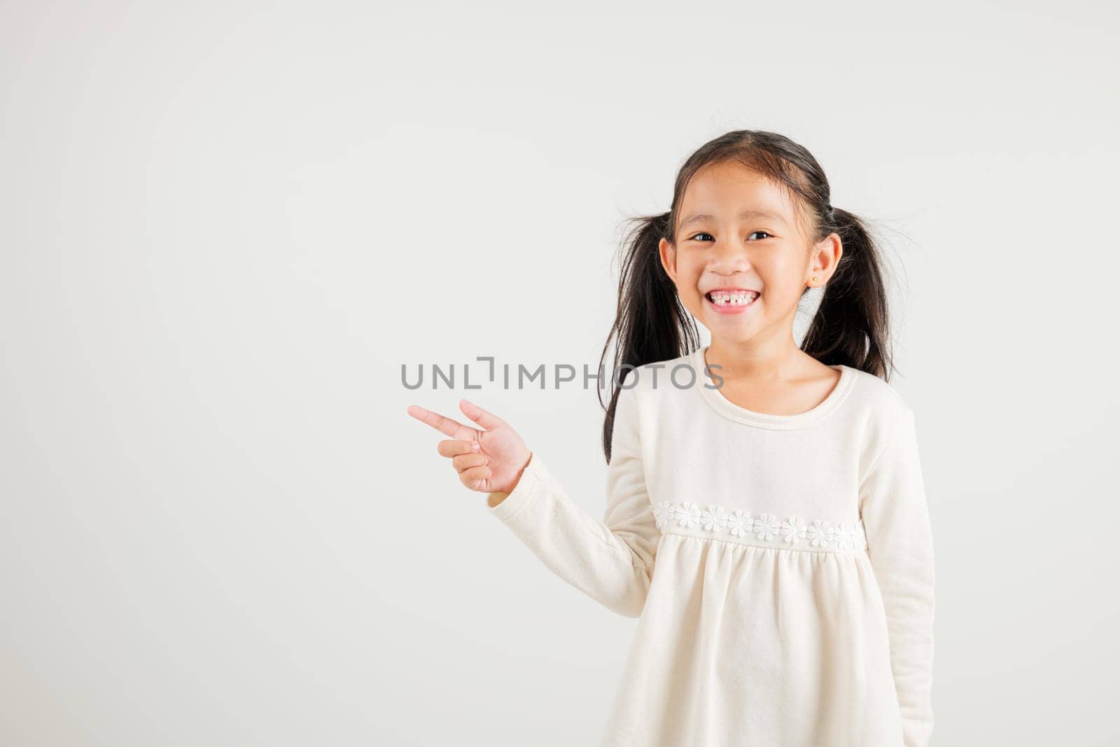 Happy Asian portrait cute young kid girl smiling indicate finger empty space, studio shot isolated on white background, Thai kindergarten child pointing index out with copy space