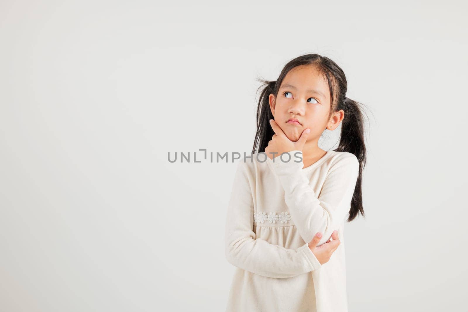 Portrait Asian kid girl deep in thought, standing with chin in hand, pondering a question. Studio shot isolated on white with ample copy space, kindergarten child portraying contemplative expression