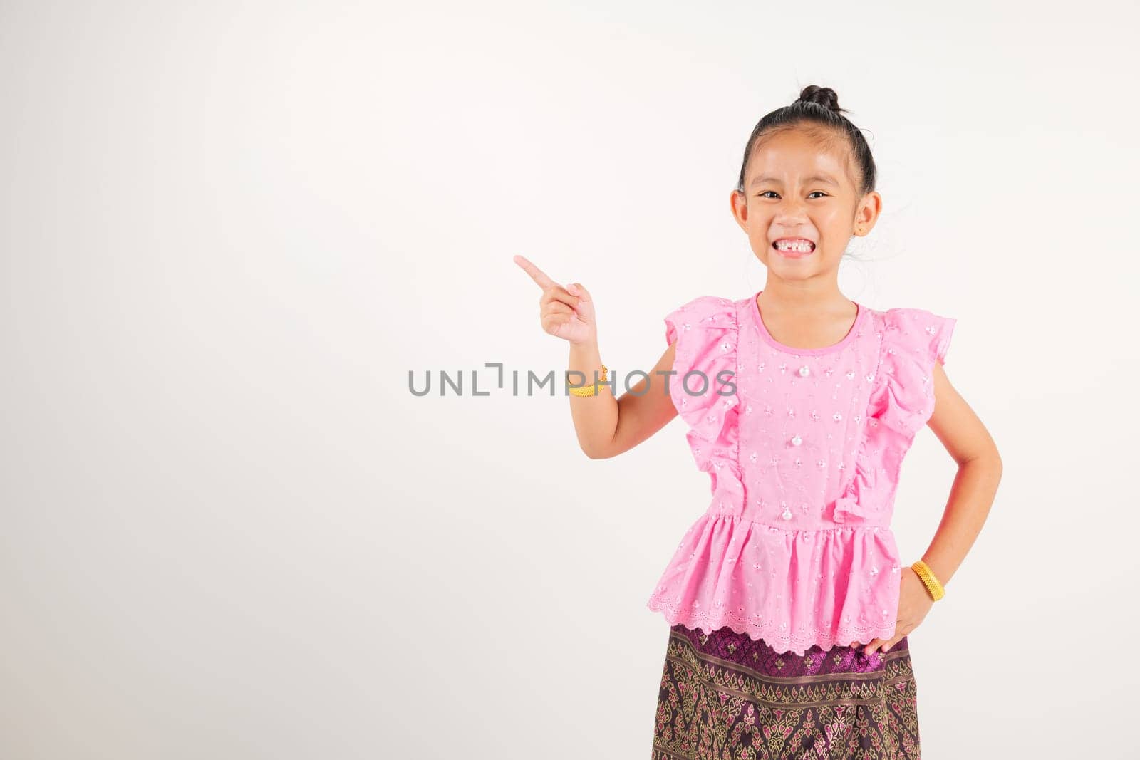 Portrait Thailand kid girl smiling traditional Thai dress costume indicate finger empty space, studio isolated white background, kindergarten child pointing index out with copy space