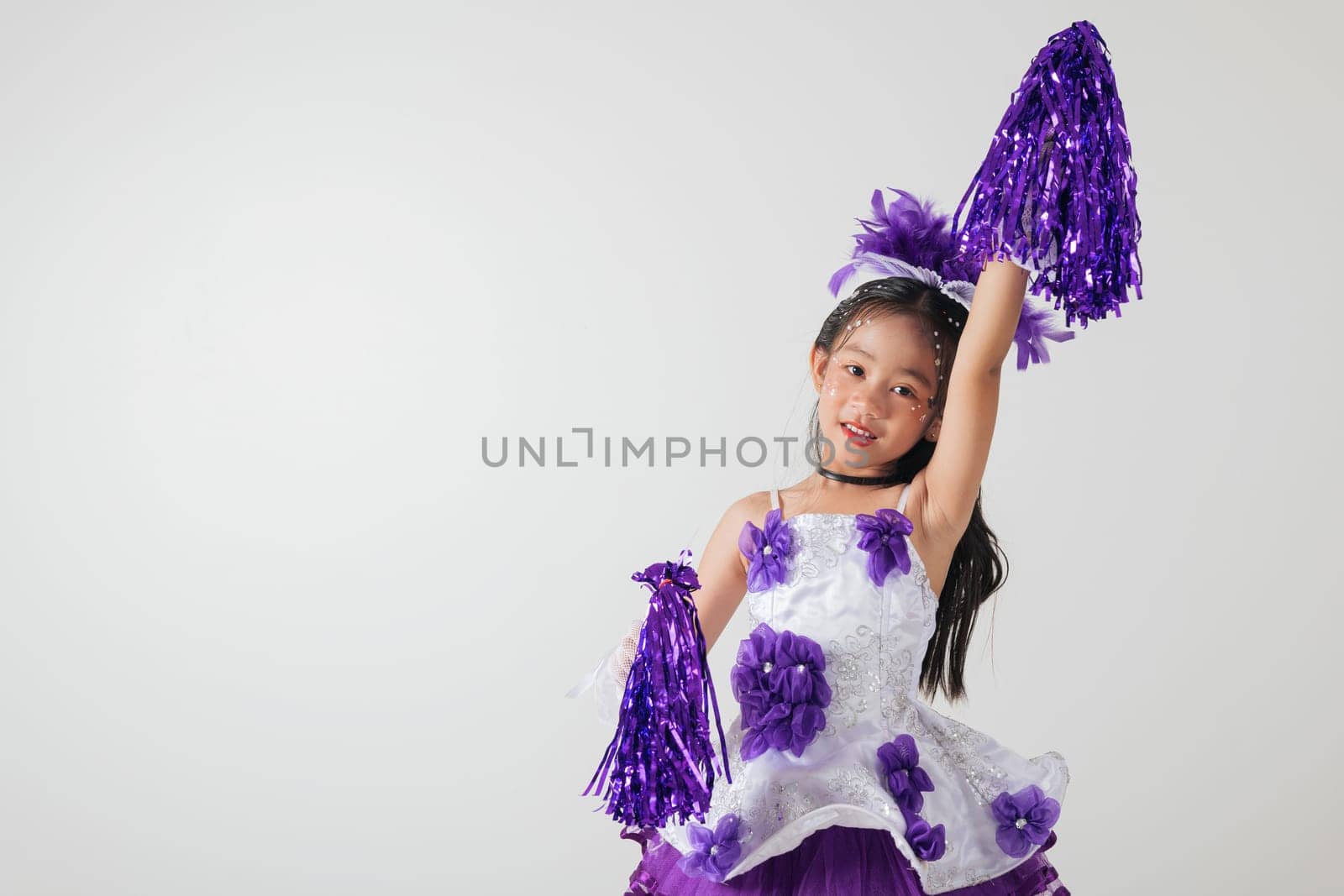 Little girl in cheerleading outfit with purple pom-poms, beaming happily in a white studio setup. A moment of fun and energy. cheerleader kid