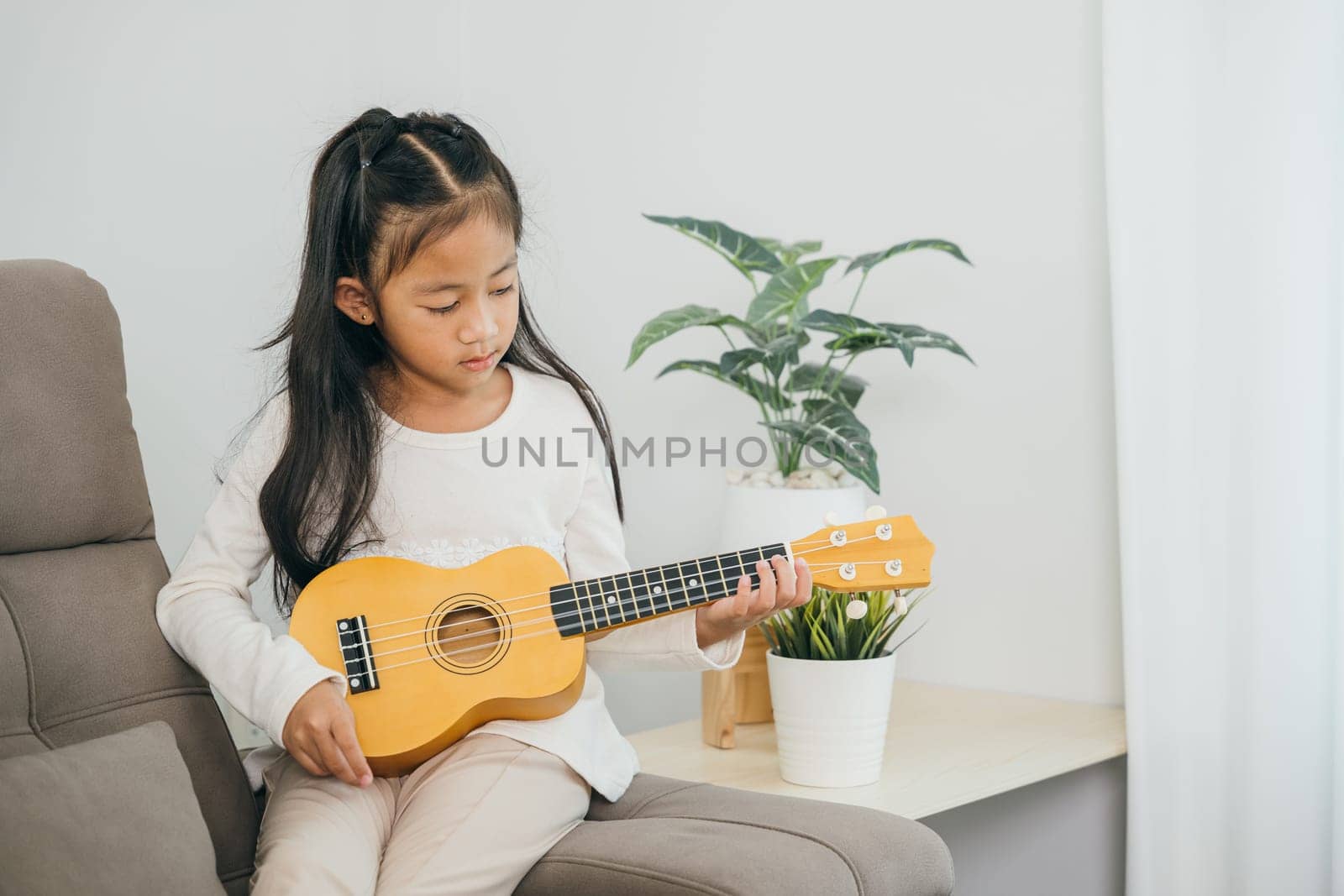 Little Asian girl plays a ukulele on a stylish sofa. Dressed in a white shirt, the living room soft tones and decor highlight the peaceful and joyful mood of this musical moment.