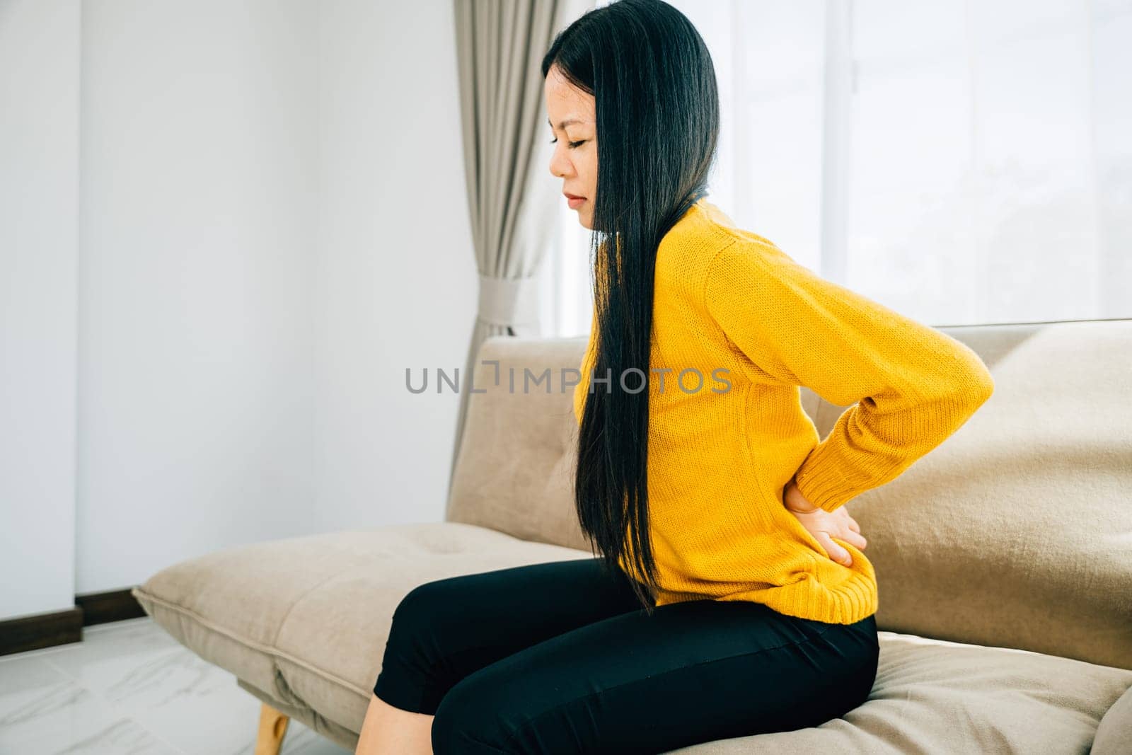 Depicting unbearable back pain, Asian woman seated on a sofa holds her lower back. Illustrating chronic backache discomfort and the necessity of medical care for back problems. health care concept