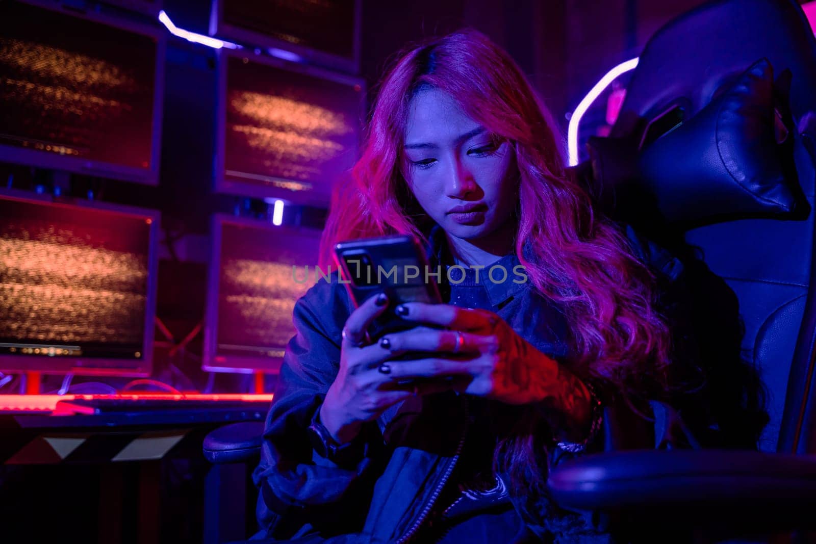 Asian woman in front of a computer monitor display using mobile phone, neon light, Hacker wear black jacket at system control room, Security guard working on multiple monitors together smartphone