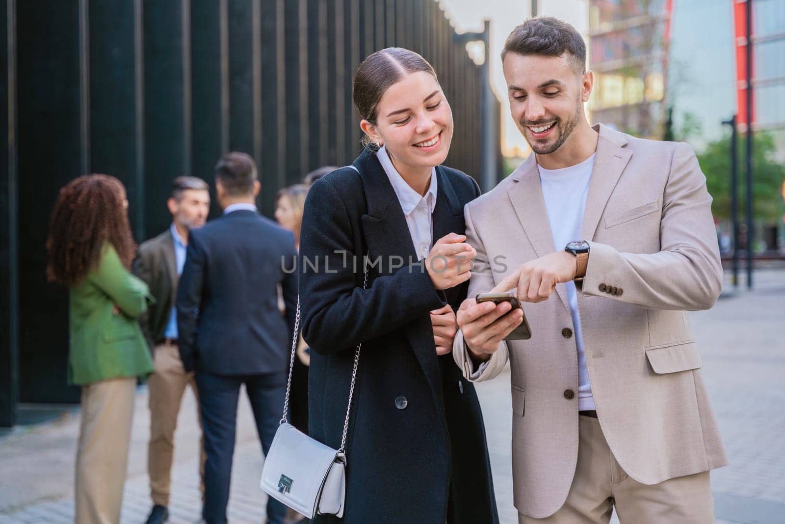 Two young business colleagues smiling while looking at a smartphone. They are standing outdoors in a modern urban setting, with other professionals in the background.
