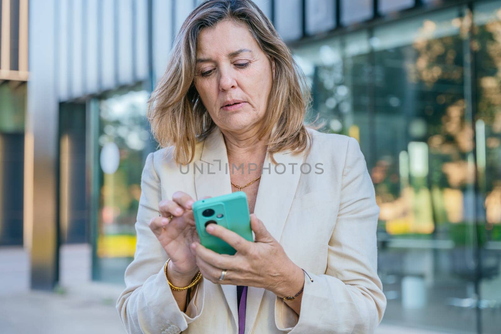 Smiling middle-aged businesswoman using a smartphone outdoors in a modern urban setting.