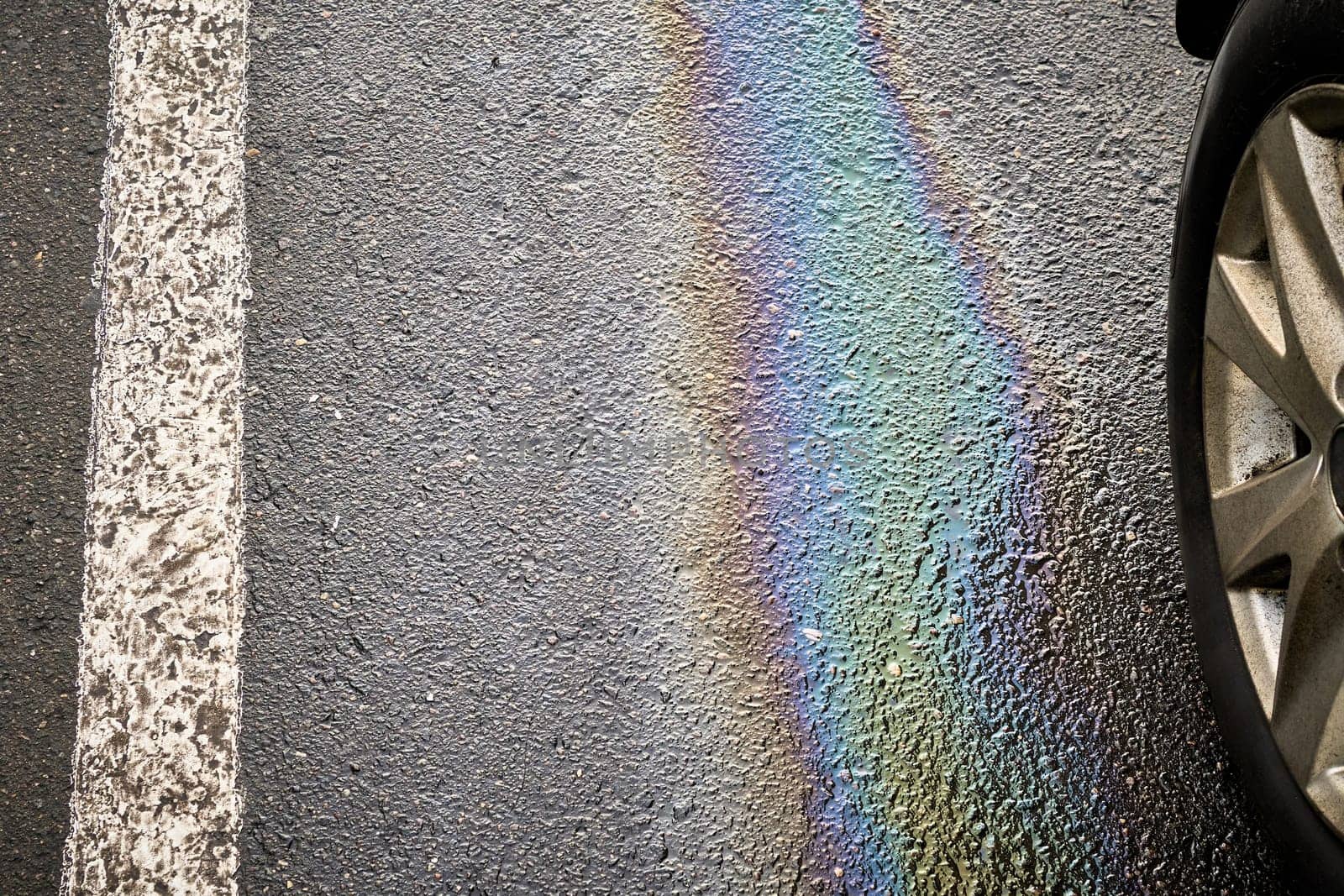 Colorful gasoline or oil spill creating a rainbow sheen on asphalt parking lot next to car tire and white parking line