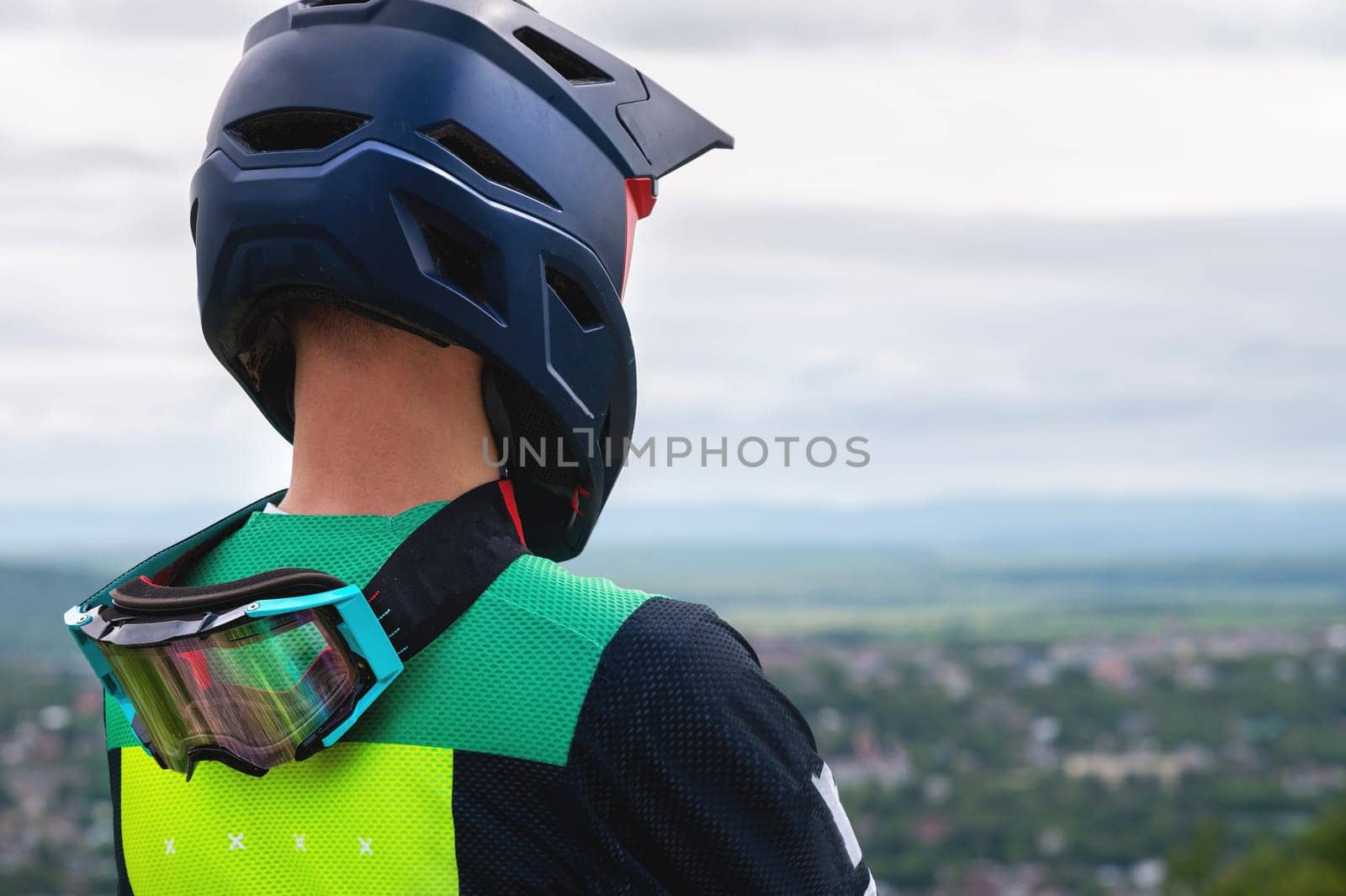 The back of a man in a protective helmet while he stands and looks at the city from the mountain. Professional racer, fully equipped with protective equipment, rests