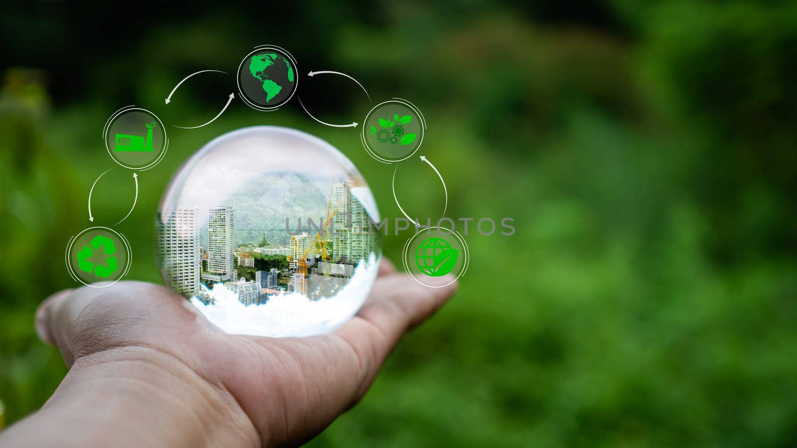 Concept of renewable energy, environmental protection, and sustainable renewable energy sources. An image of a building under construction in crystal glass resting on a person's hand and with a natural green background. Clean energy.