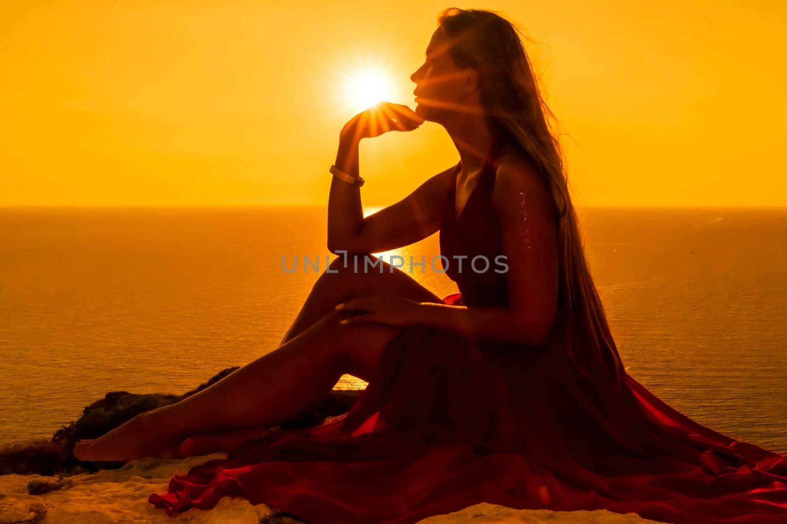 Woman sunset sea red dress, side view a happy beautiful sensual woman in a red long dress posing on a rock high above the sea on sunset