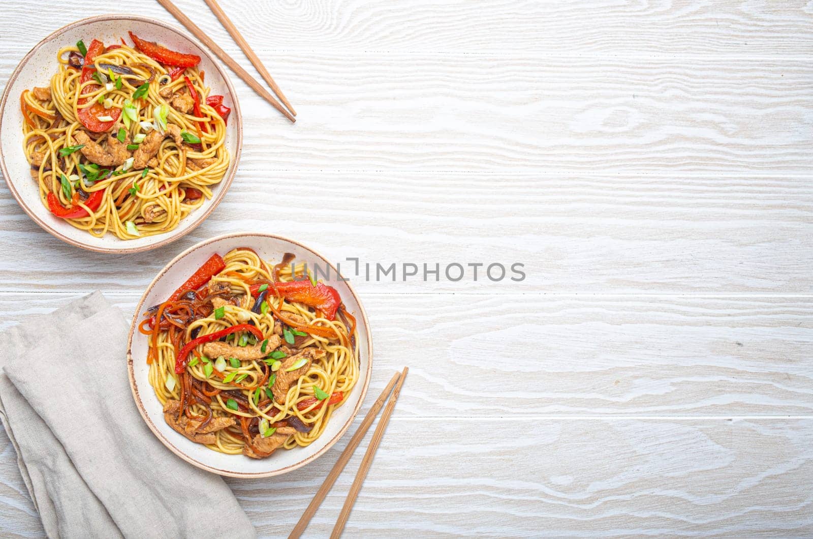 Two bowls with Chow Mein or Lo Mein, traditional Chinese stir fry noodles with meat and vegetables, served with chopsticks top view on rustic white wooden background table, space for text.