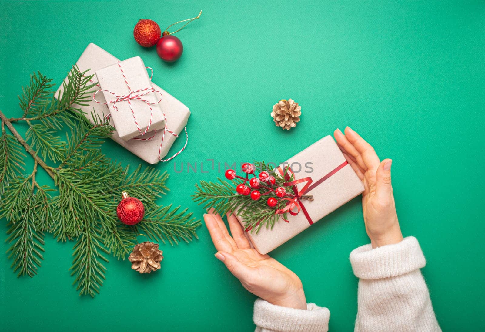 Christmas or New Year celebration green paper festive background with female hands holding wrapped gift box, decoration fir tree, cones, berries, sparkly red balls. Giving presents concept. by its_al_dente