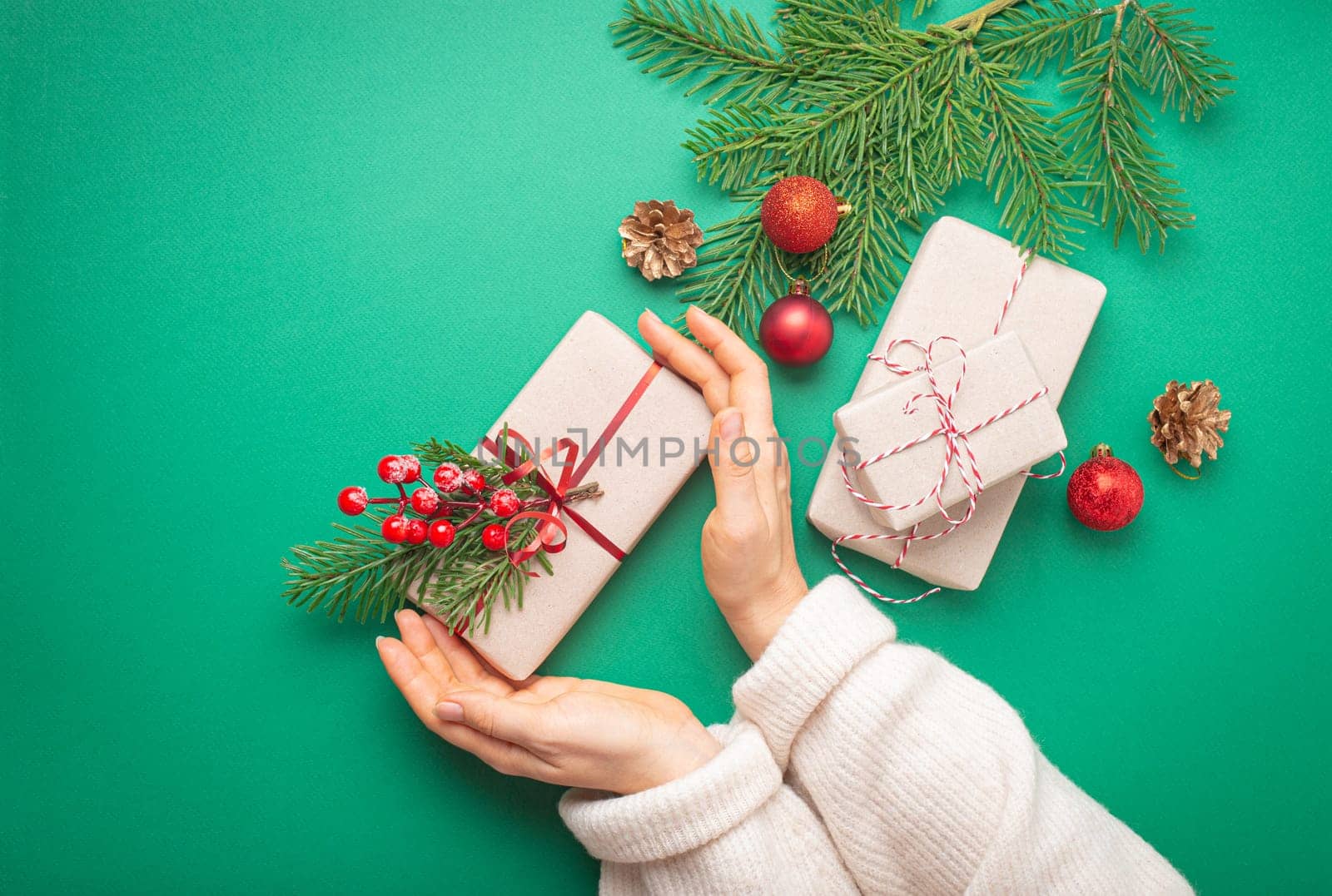 Christmas or New Year celebration green paper festive background with female hands holding wrapped present box, decoration fir tree, cones, berries, sparkly red balls. Space for text..