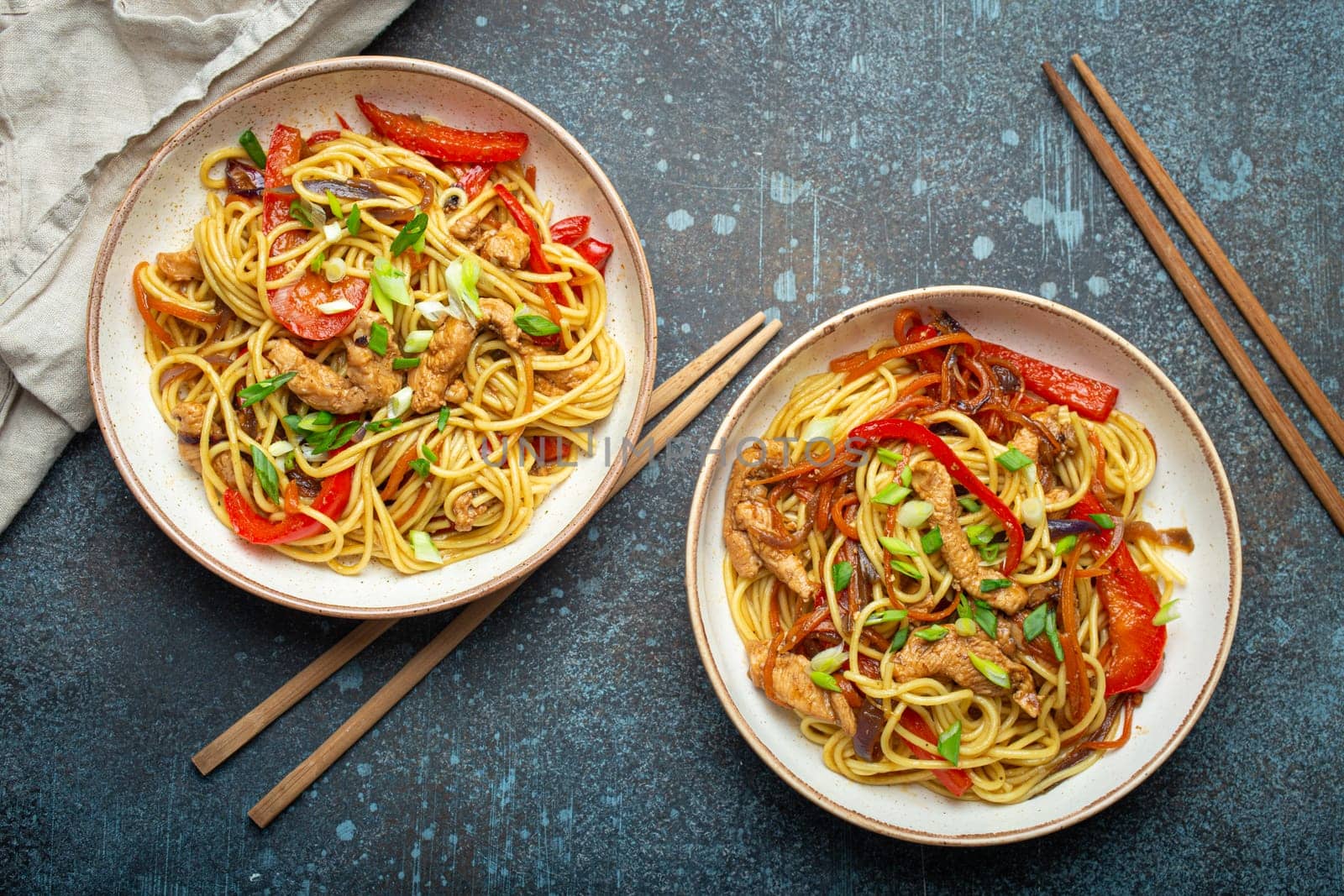 Two bowls with Chow Mein or Lo Mein, traditional Chinese stir fry noodles with meat and vegetables, served with chopsticks top view on rustic blue concrete background.