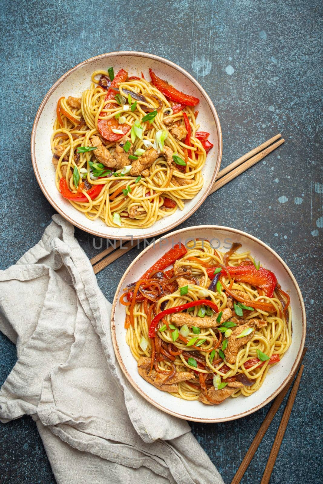 Two bowls with Chow Mein or Lo Mein, traditional Chinese stir fry noodles with meat and vegetables, served with chopsticks top view on rustic blue concrete background.