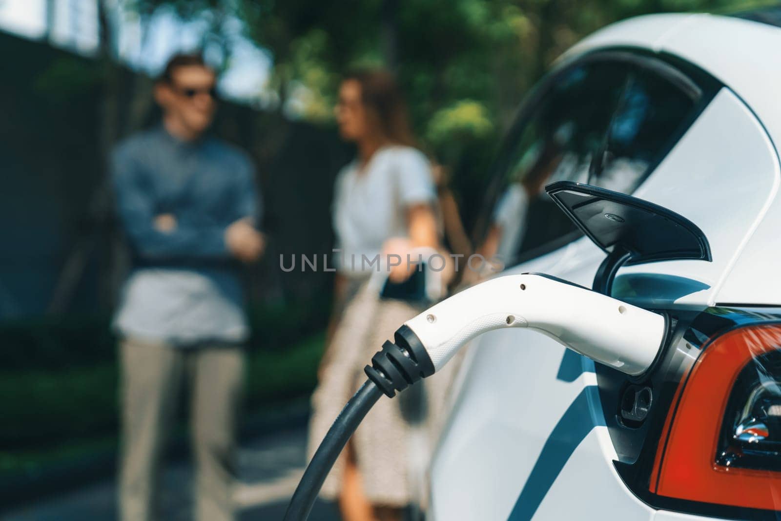 Young couple travel with EV electric car charging in green sustainable city outdoor garden in summer shows urban sustainability lifestyle by green clean rechargeable energy of electric vehicle innards