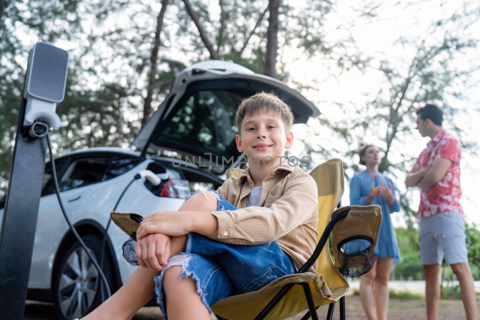 Little boy portrait sitting on camping chair with his family in background. Road trip travel with alternative energy charging station for eco-friendly car concept. Perpetual