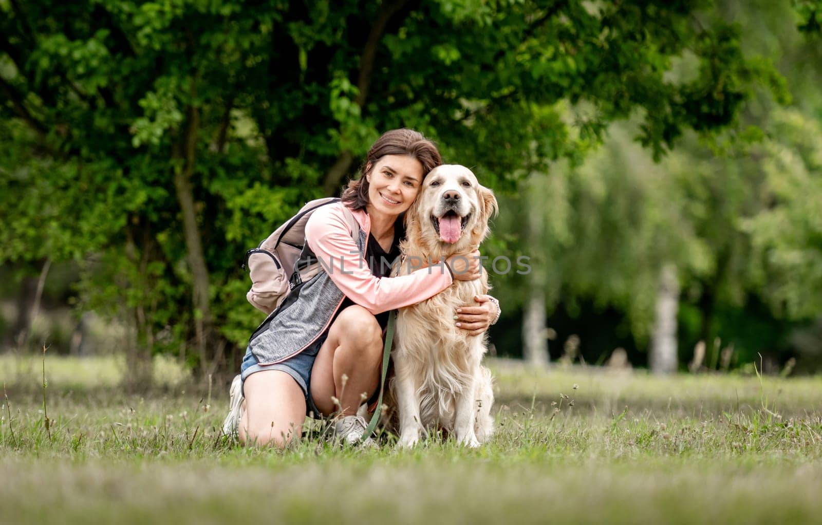 Pretty girl with golden retriever dog sitting at nature. Beautiful young woman hugging purebred pet doggy labrador in park at summer