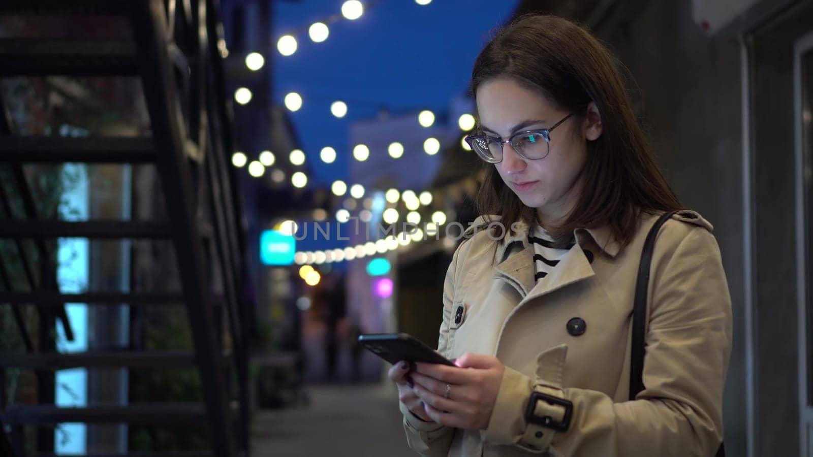 A young woman chats on a smartphone late at night on a narrow street. A girl in glasses with a phone in hands. 4k