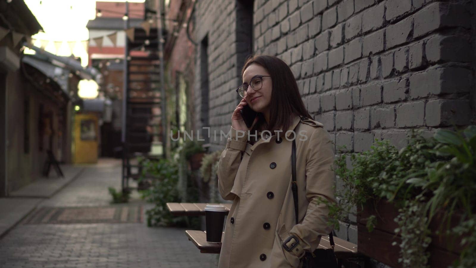 A young woman talks on the phone while standing against a wall on a narrow street. A girl with glasses and a coat speaks on the phone. 4k