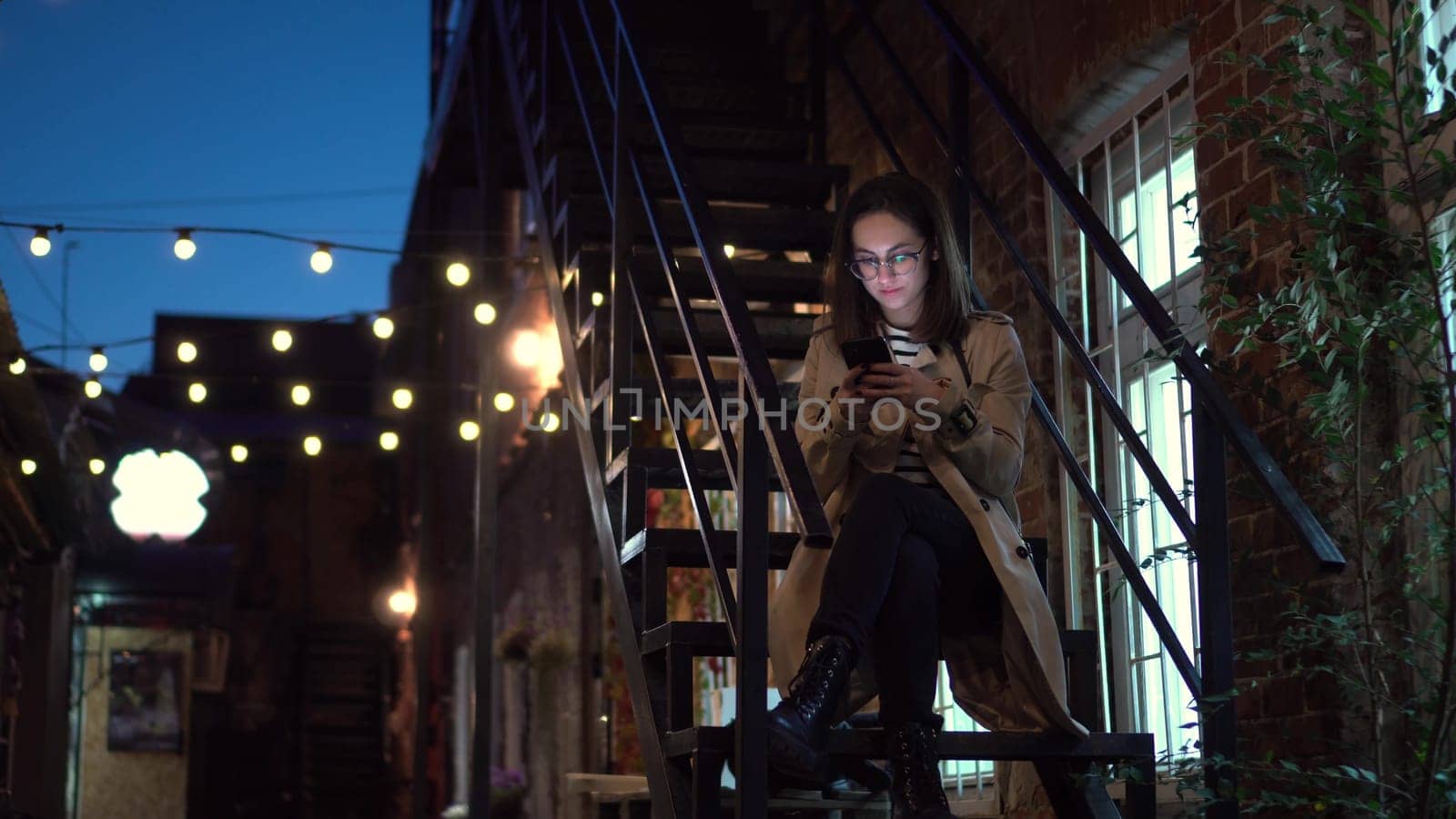 A young woman sits on a fire escape and texts on a smartphone. A girl with glasses on a narrow street late at night with a phone in her hands. 4k