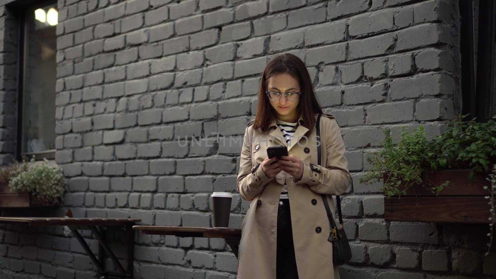 A young woman is chatting on a smartphone while standing against a wall on a narrow street. A girl in glasses and a polo with a phone in her hands. 4k