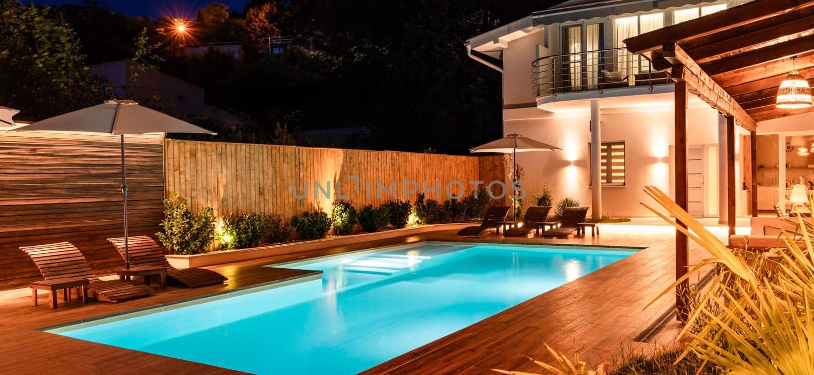 Luxury backyard with illuminated swimming pool and modern house. Elegant outdoor space featuring wooden deck and cozy ambiance