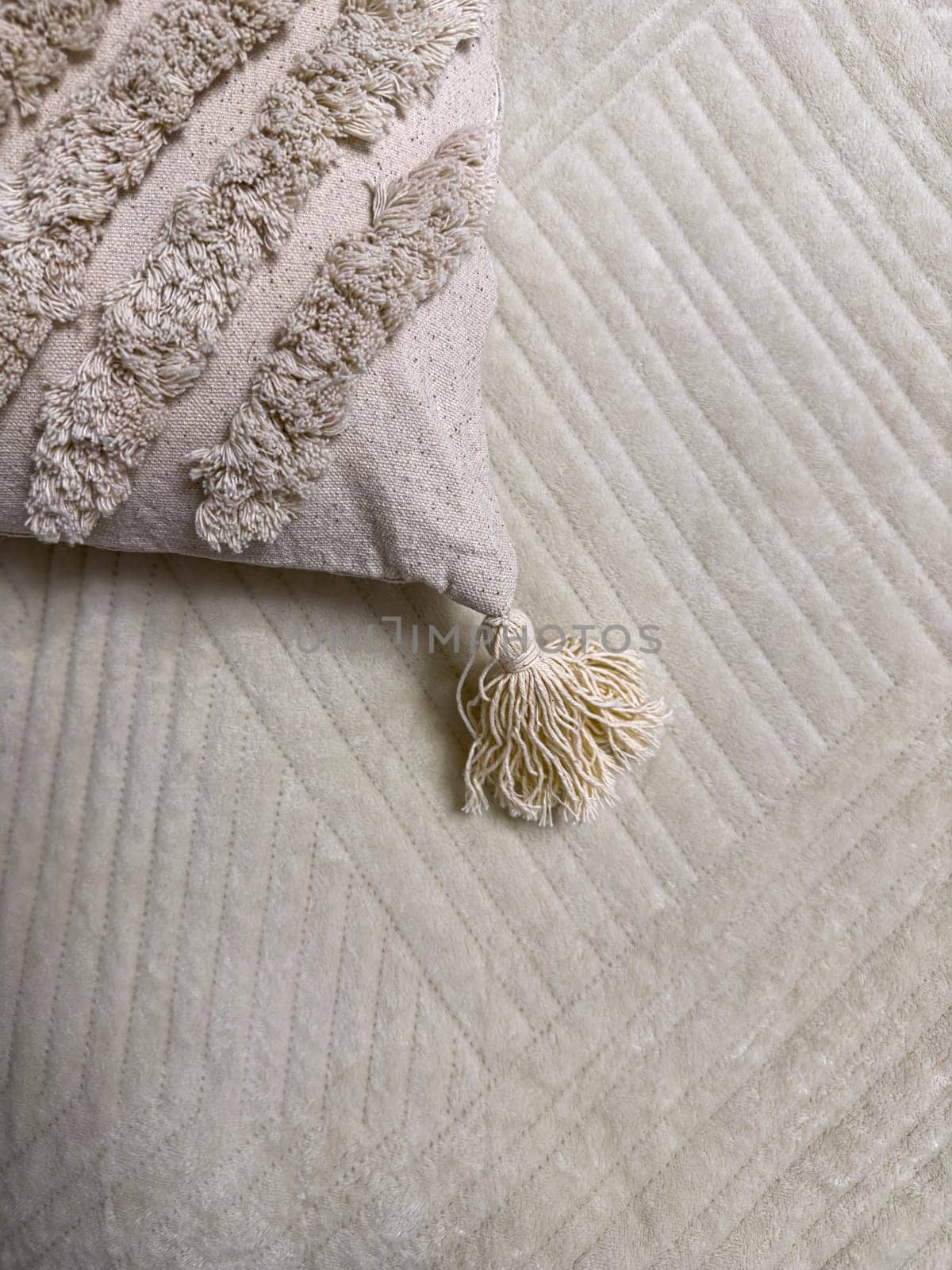 A soft, textured pillow with a tassel sits atop a light-colored quilted blanket. The neutral tones create a cozy and inviting atmosphere, perfect for relaxation.