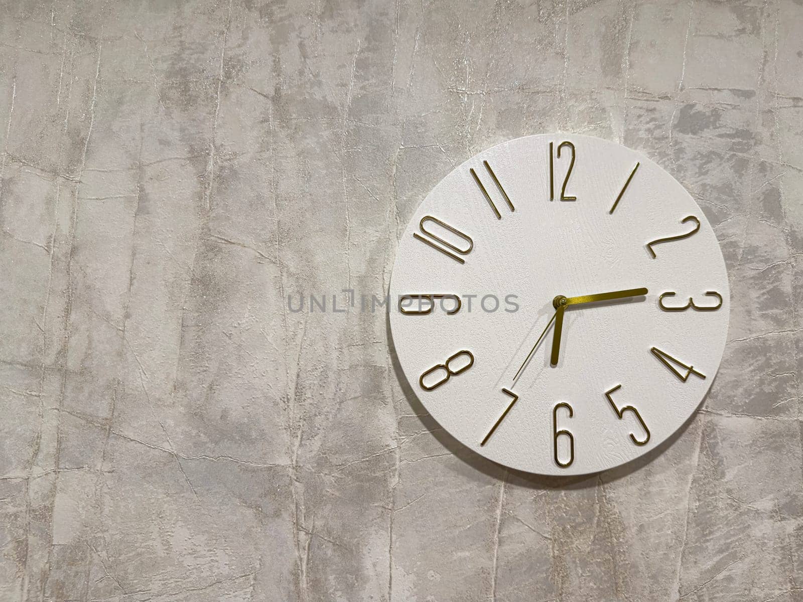 A stylish white wall clock featuring large black numbers is mounted on a textured gray wall. The clock has a minimalist design, showcasing a simple yet elegant look.