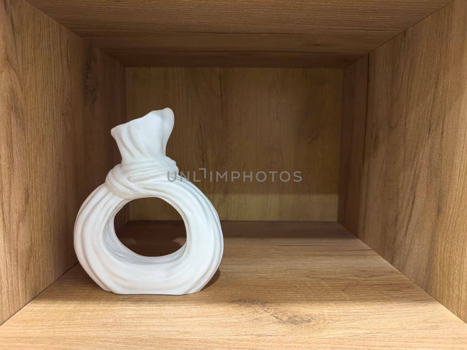 A white ceramic vase with a distinct shape sits gracefully on a natural wooden shelf. The minimalist design enhances the contemporary decor of the room, providing a stylish accent.