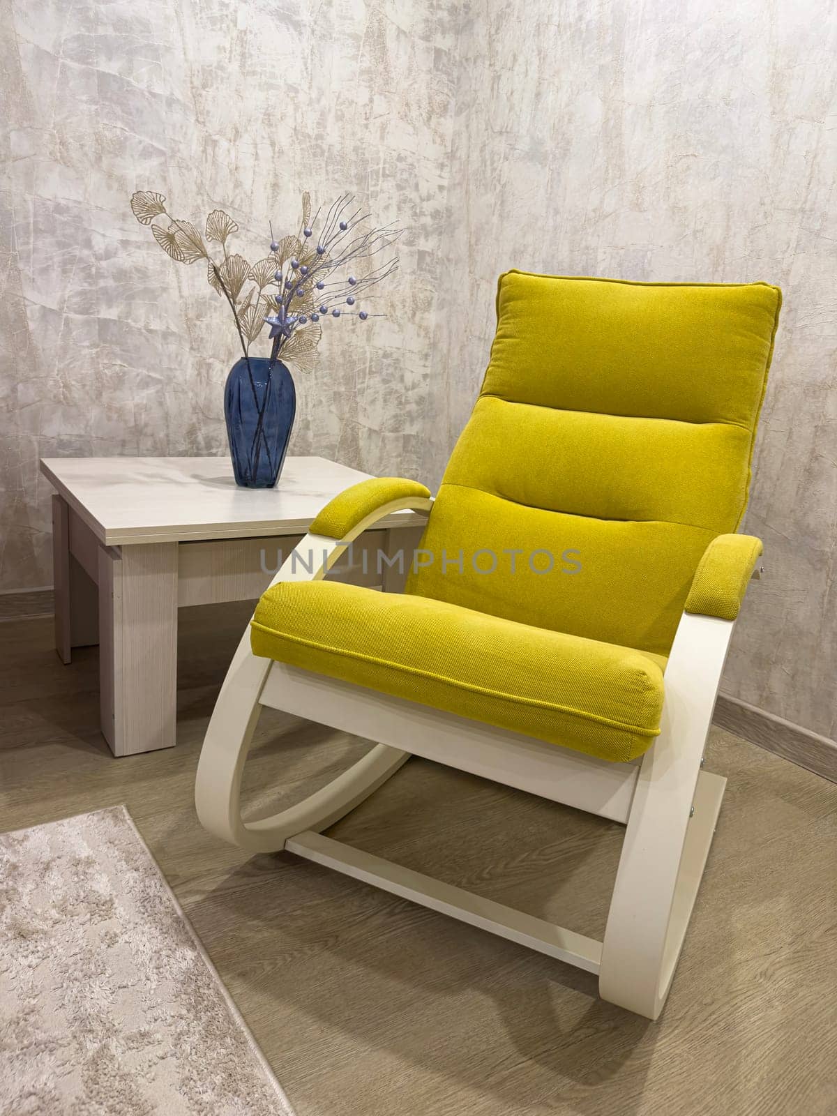 A vibrant yellow rocking chair sits next to a sleek side table adorned with a blue vase holding dried flowers, enhancing the contemporary decor of the cozy room.