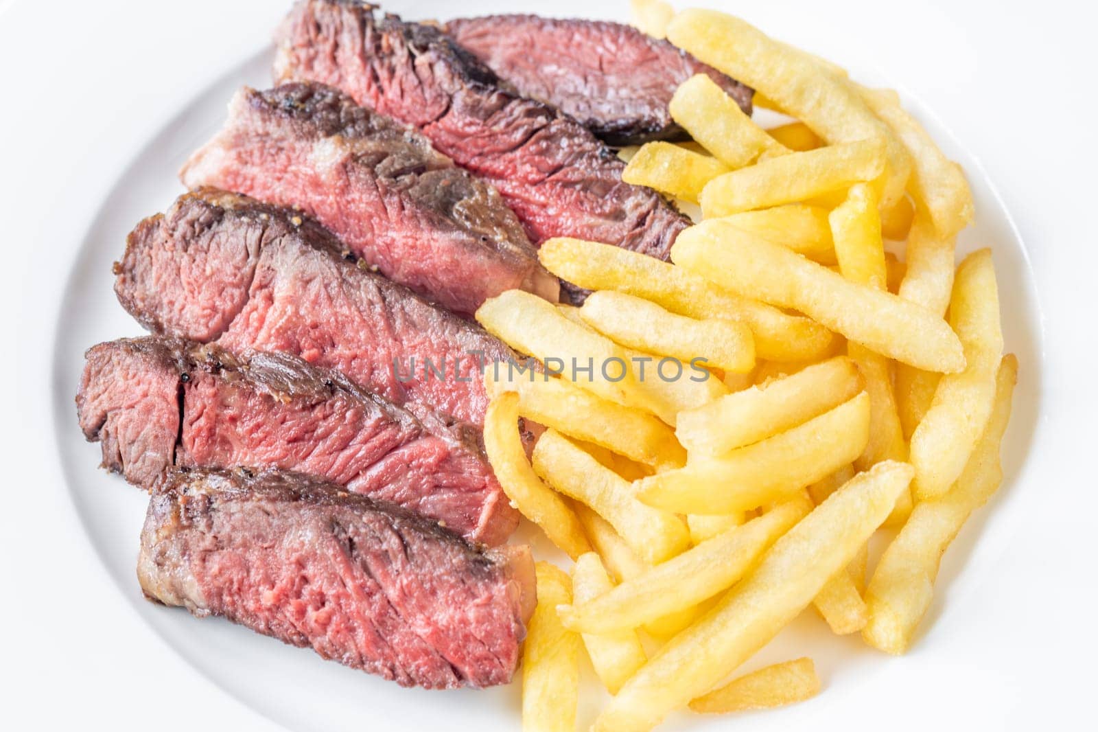 Sliced medium-rare ribeye steak with golden french fries served on a white plate on a wooden table
