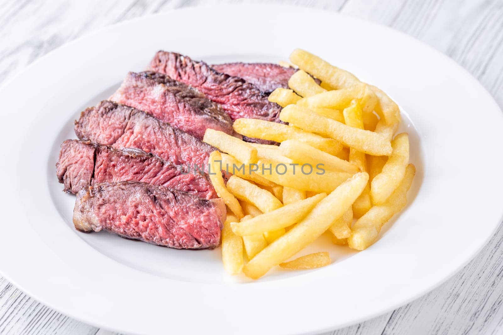 Sliced medium-rare ribeye steak with golden french fries served on a white plate on a wooden table