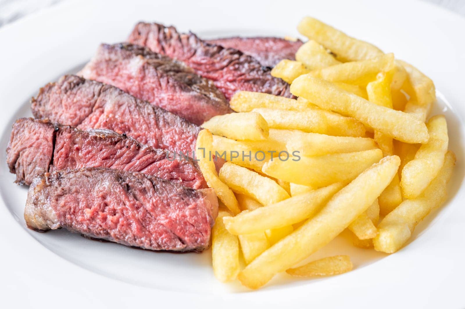 Sliced medium-rare ribeye steak with golden french fries served on a white plate on a wooden table
