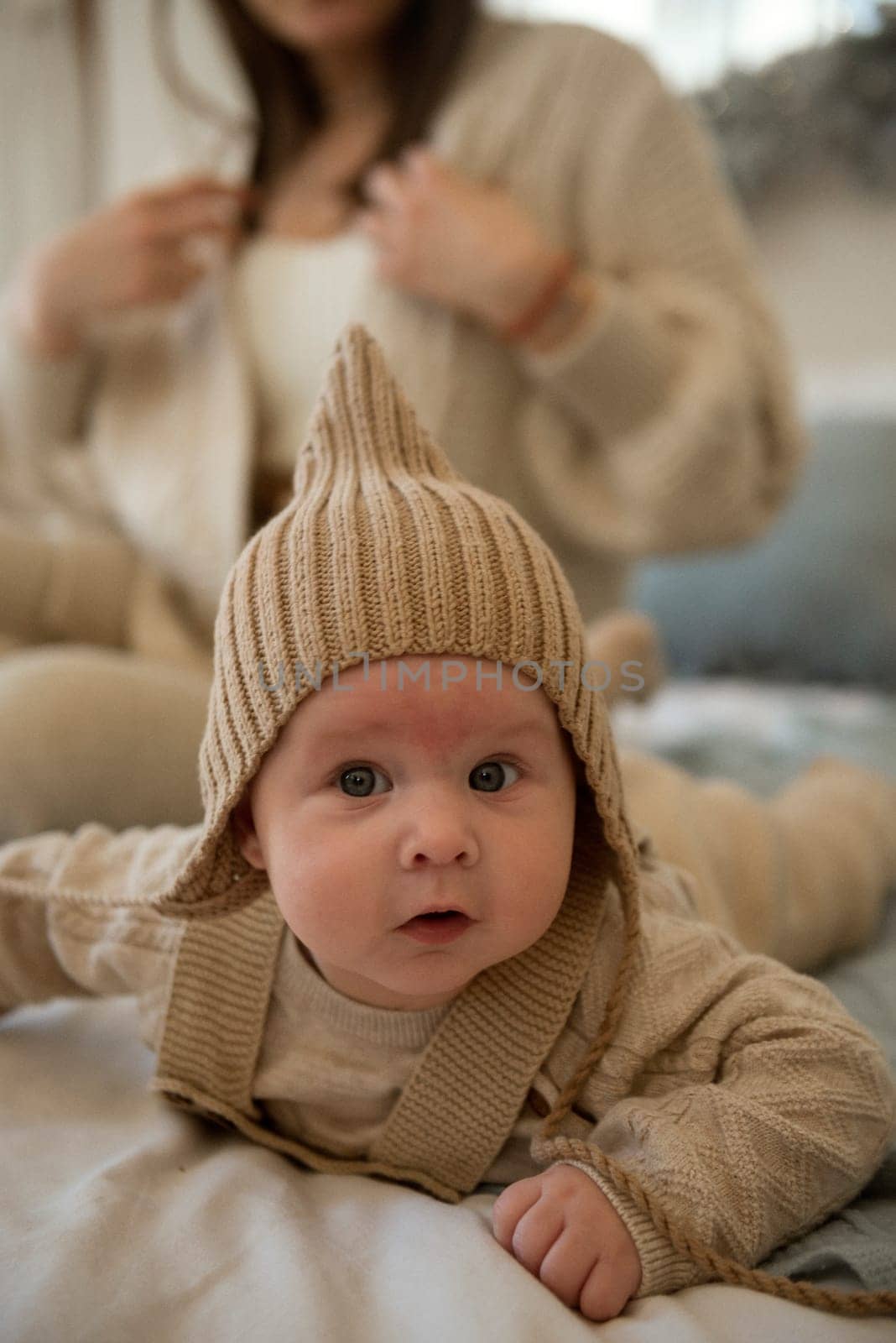A baby wearing a hat. A small child is crawling on a bed. His mother is sitting behind him. The little boy is wearing a knitted beige hat. the baby is very active and cute, he crawls on the bed.
