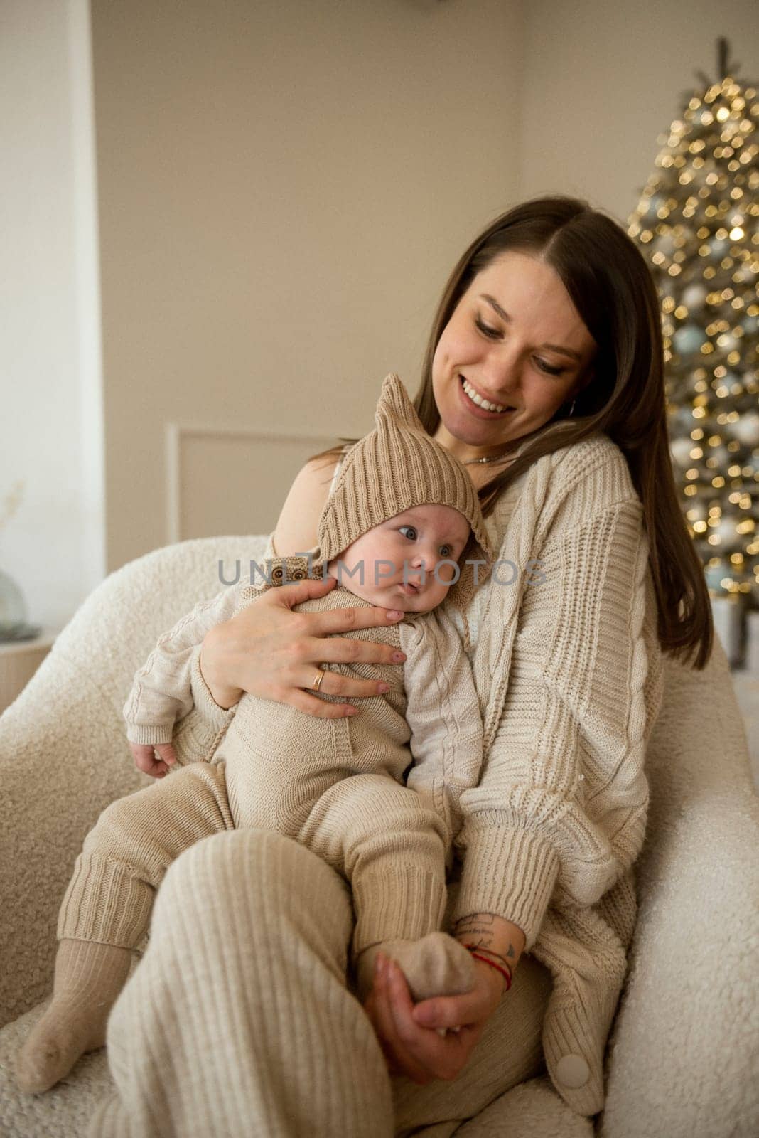 A woman holding a baby.The girl holds her child in her arms. The child is wearing a beige hand-knitted hat. mother and child are happy, they are smiling. They celebrate Christmas.