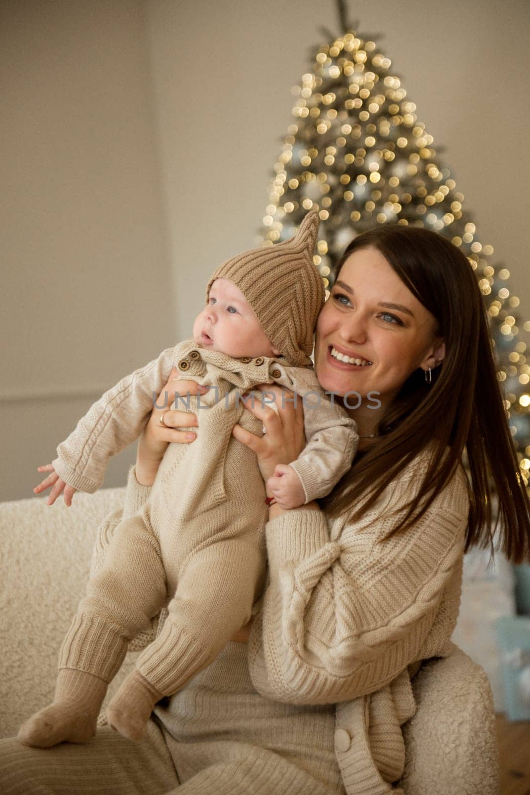 A woman holding a baby.The girl holds her child in her arms. The child is wearing a beige hand-knitted hat. mother and child are happy, they are smiling. They celebrate Christmas.