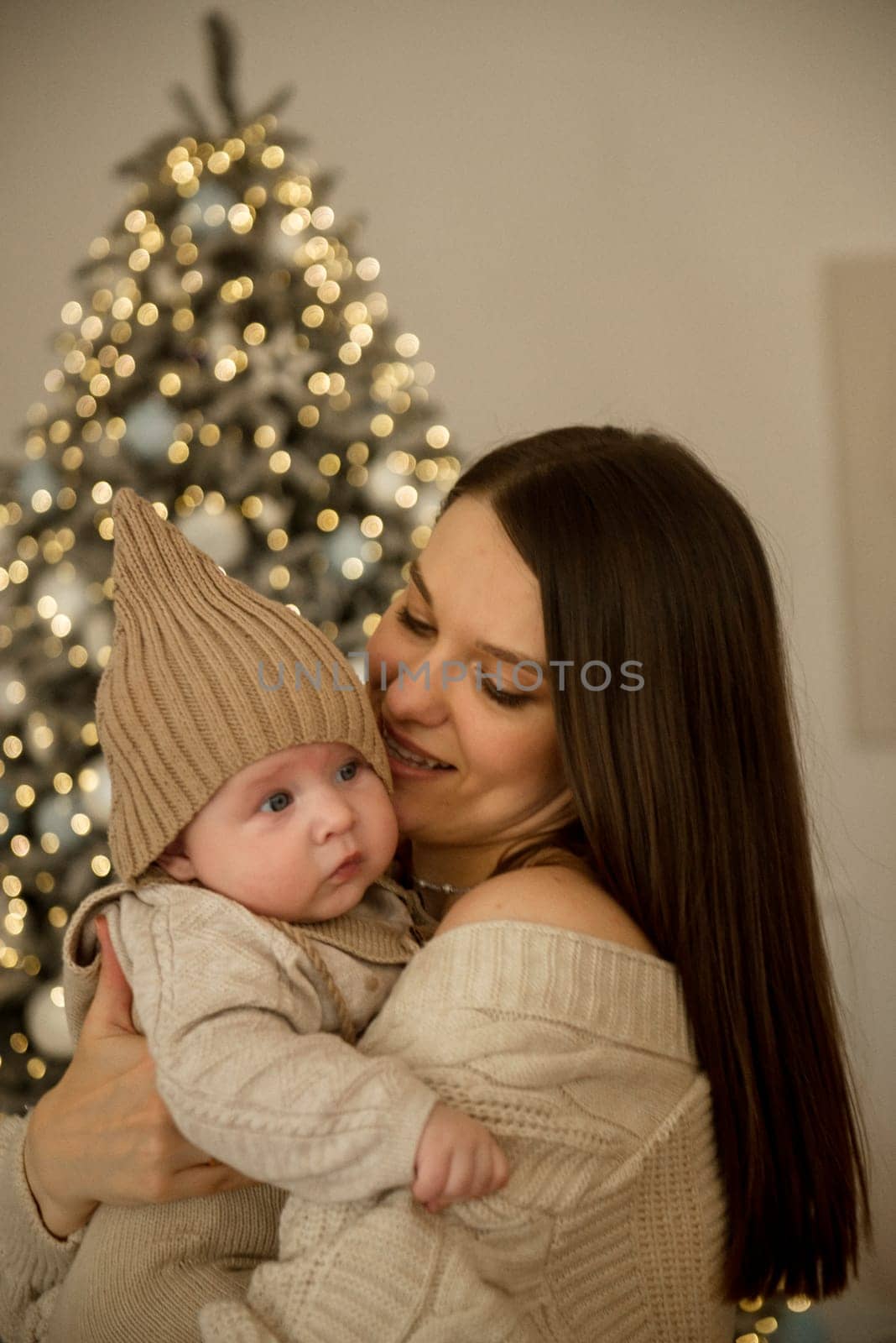 A woman holding a baby.The girl holds her child in her arms. The child is wearing a beige hand-knitted hat. mother and child are happy, they are smiling. They celebrate Christmas.