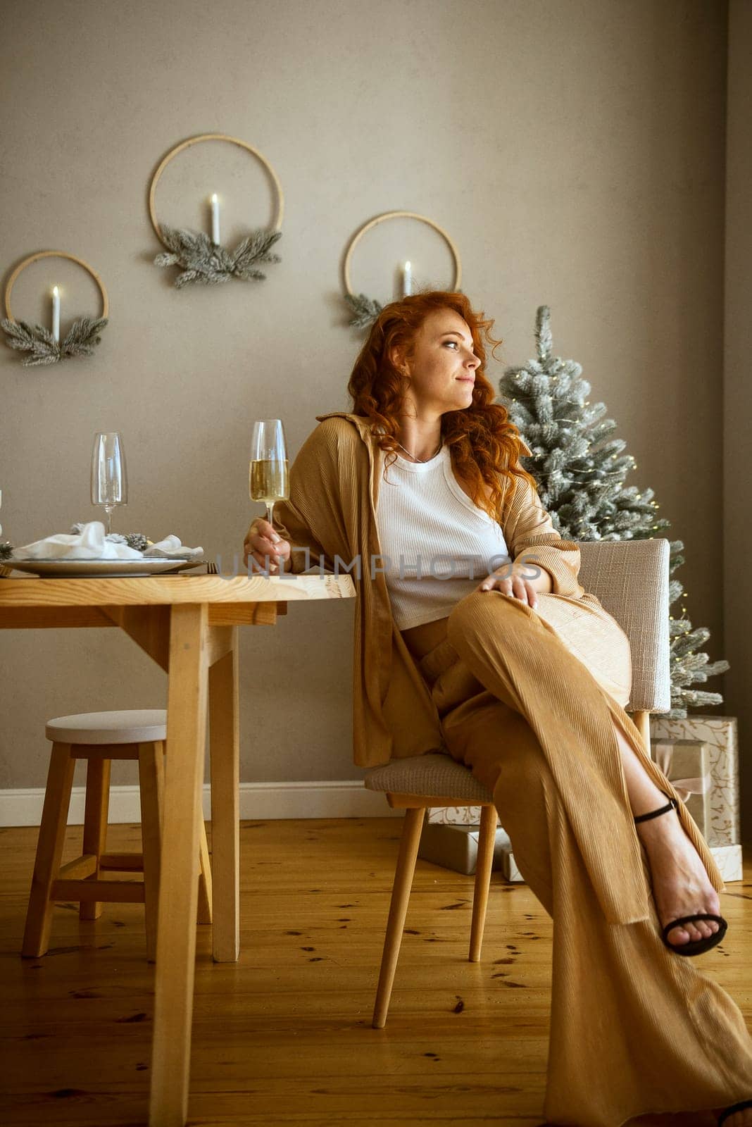 A person sitting on a table. The girl is sitting in the room at the table. She has long red hair. There is Christmas decor behind her. The girl is smiling, She has a wine glass in her hands