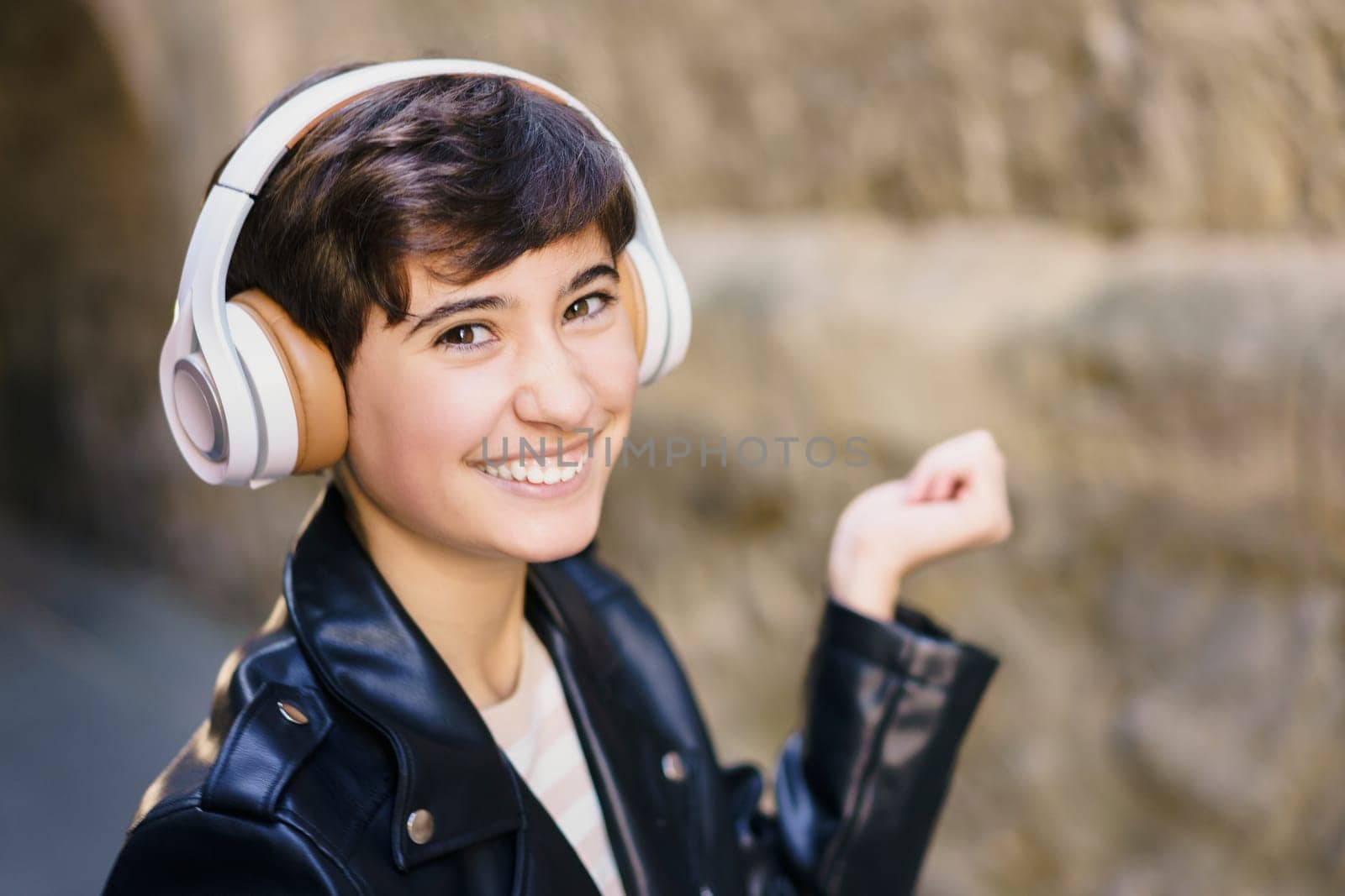 A cheerful young person is wearing trendy headphones while smiling broadly and making cheerful gestures in a vibrant urban setting, illustrating a deep passion for music and a carefree lifestyle