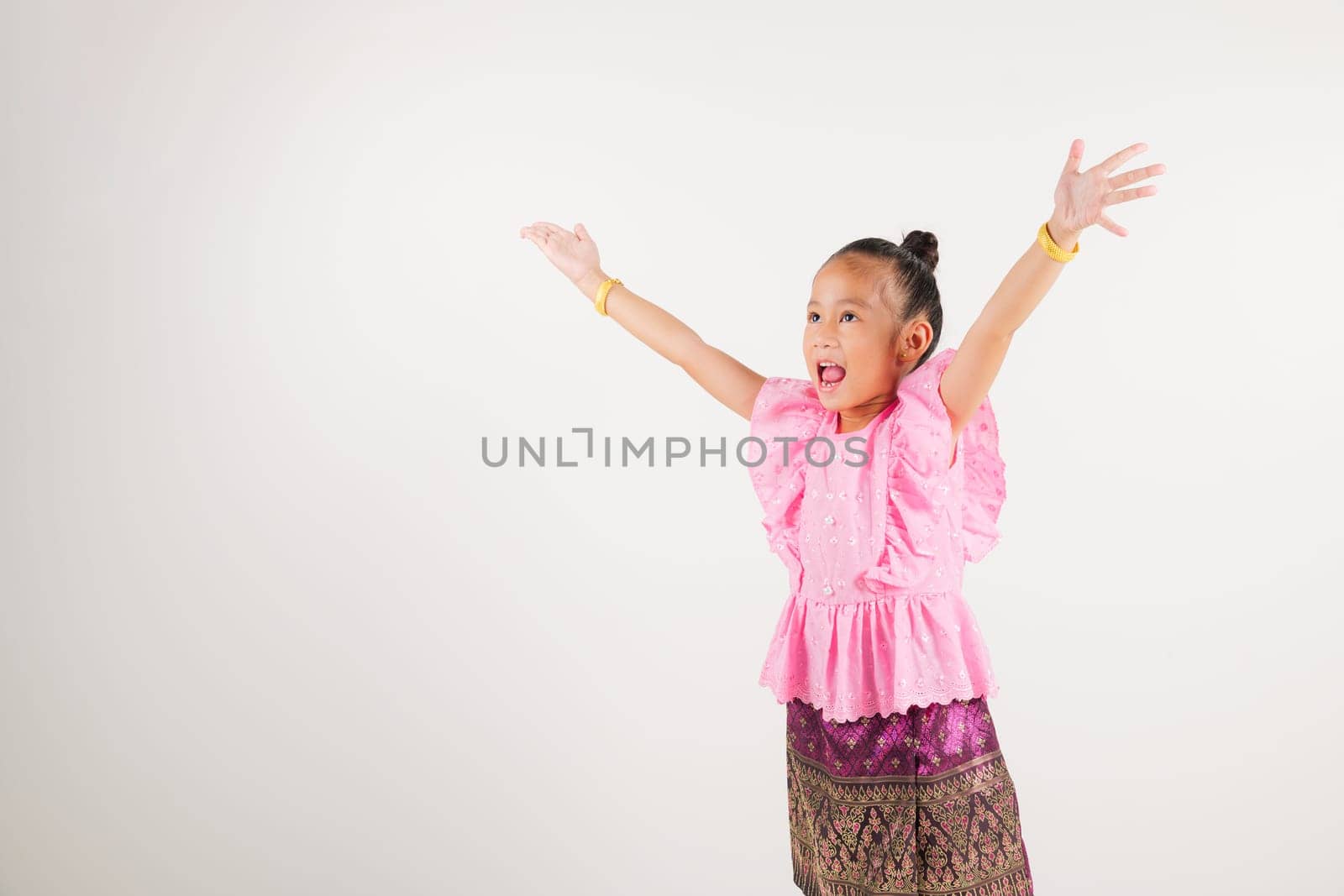 Portrait Thailand kid girl smiling traditional Thai dress costume raises fists in celebration of her success, studio isolated white background, kindergarten child saying yes with excitement winning