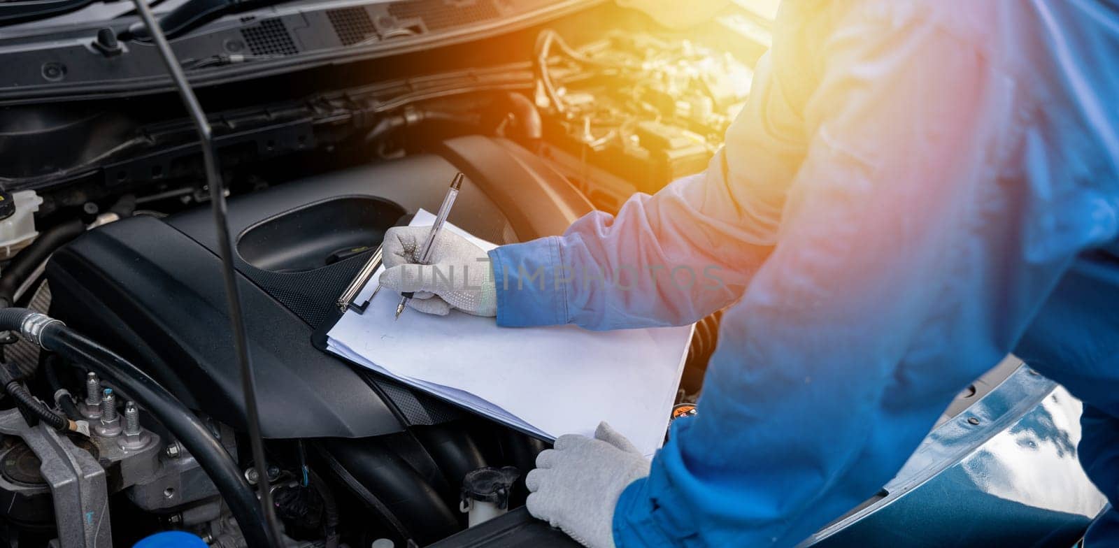 Skilled Asian mechanic in blue shirt writing on a paper while working on a car in an auto repair shop. Close-up shot of hand.