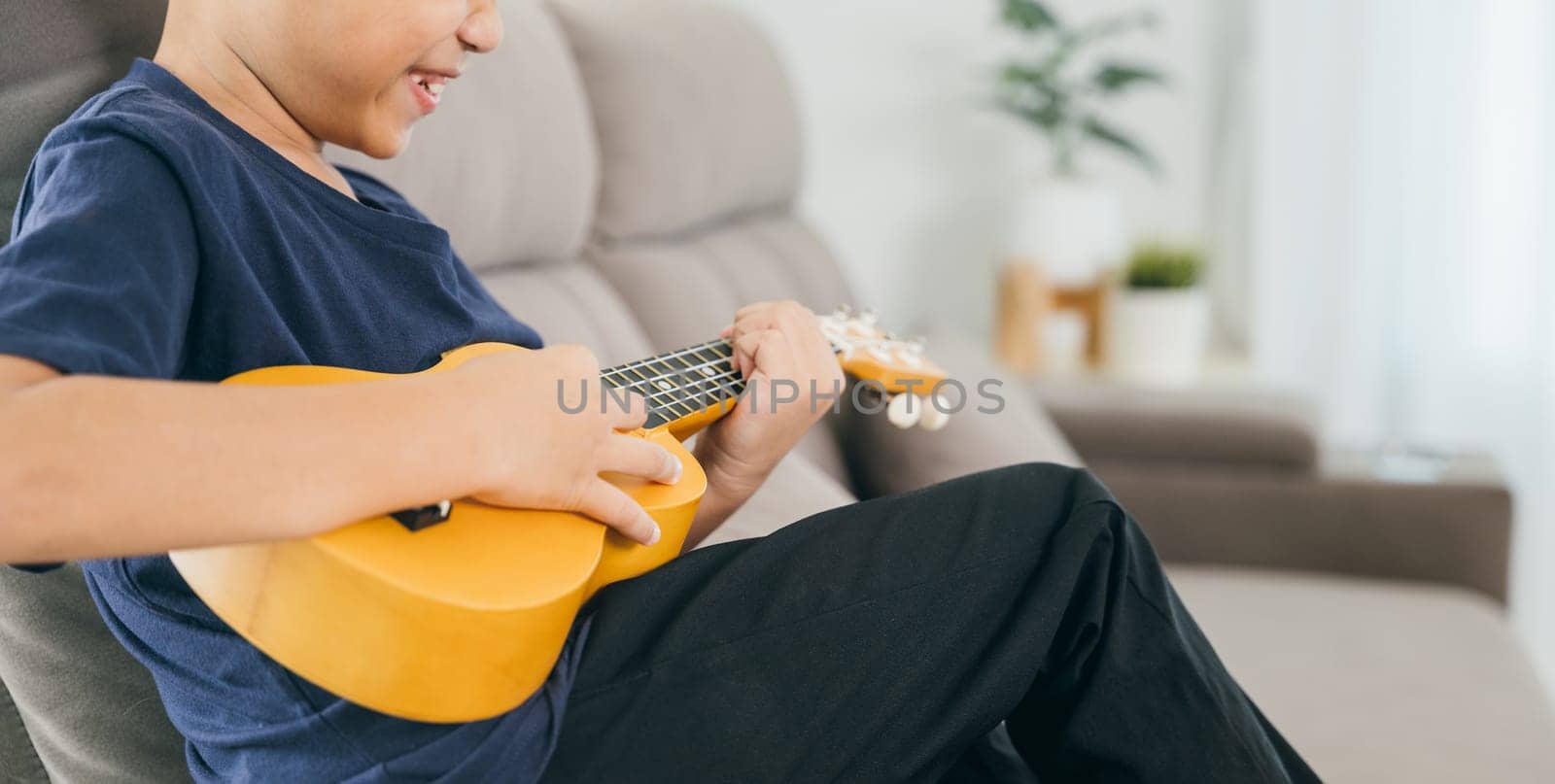 Little boy's hand strums the strings of a yellow ukulele on a couch. The cozy living room scene captures a moment of creativity and playful learning.