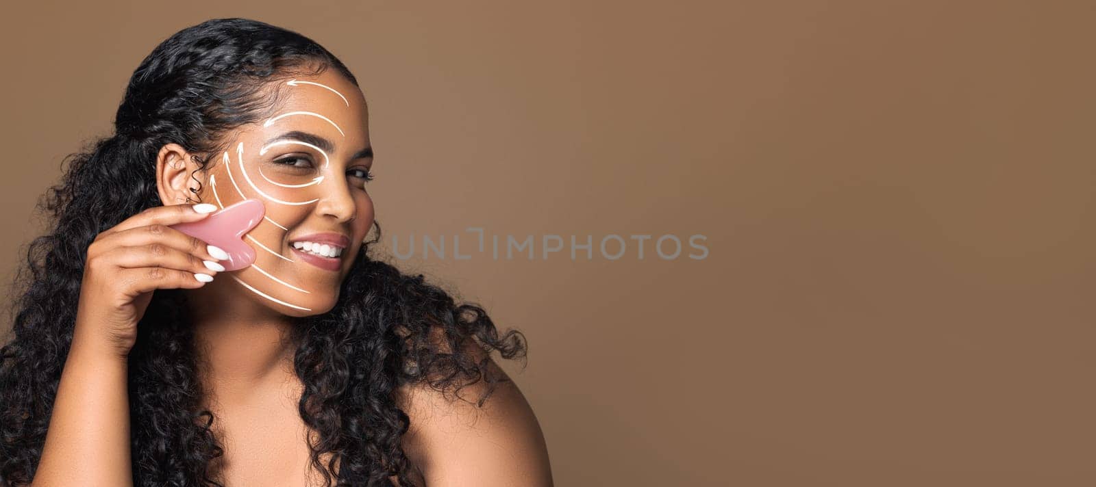 A woman enjoys her beauty routine, using a tool to apply skin care treatment on her smiling face. She is isolated against a soft brown background, emphasizing her relaxation.