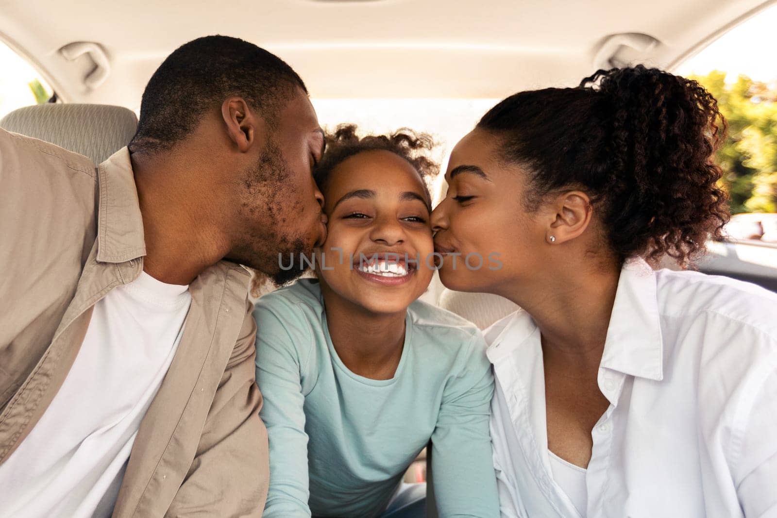 Cheerful Black Parents Kissing Daughter Sitting In New Car Riding Enjoying Road Trip Together. African American Family Having Summer Vacation And Traveling By Auto. Transportation Concept
