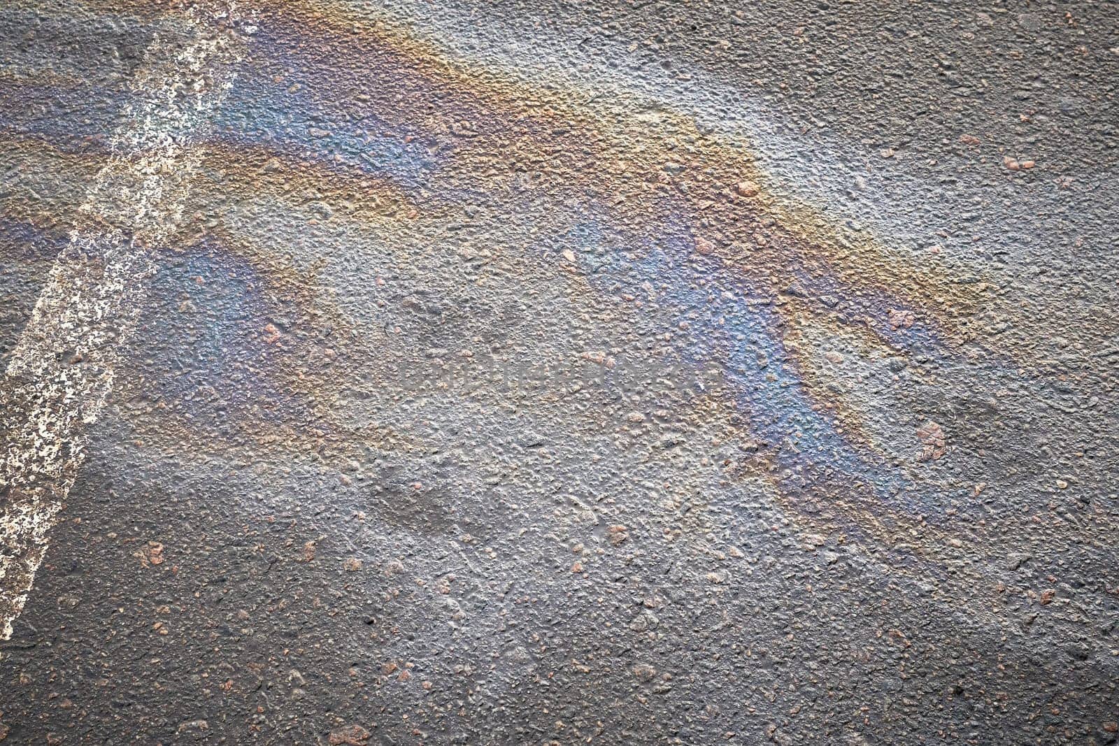 A vibrant oil spill spreads across the asphalt surface, reflecting a range of colors, contrasting with the white parking line. The scene captures an urban environment, highlighting pollution