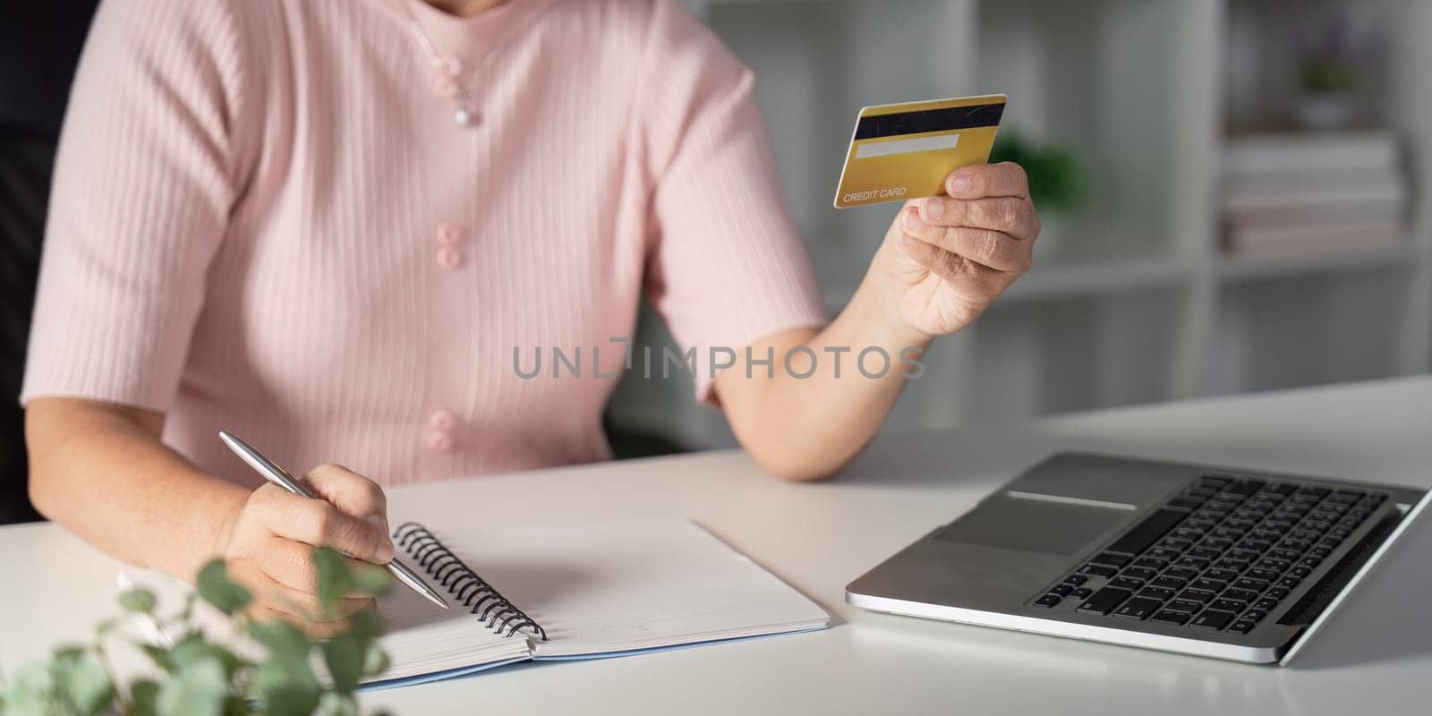 Focused elderly woman writing in her notebook while holding a credit card, preparing for her online shopping session from the comfort of her stylish home office.