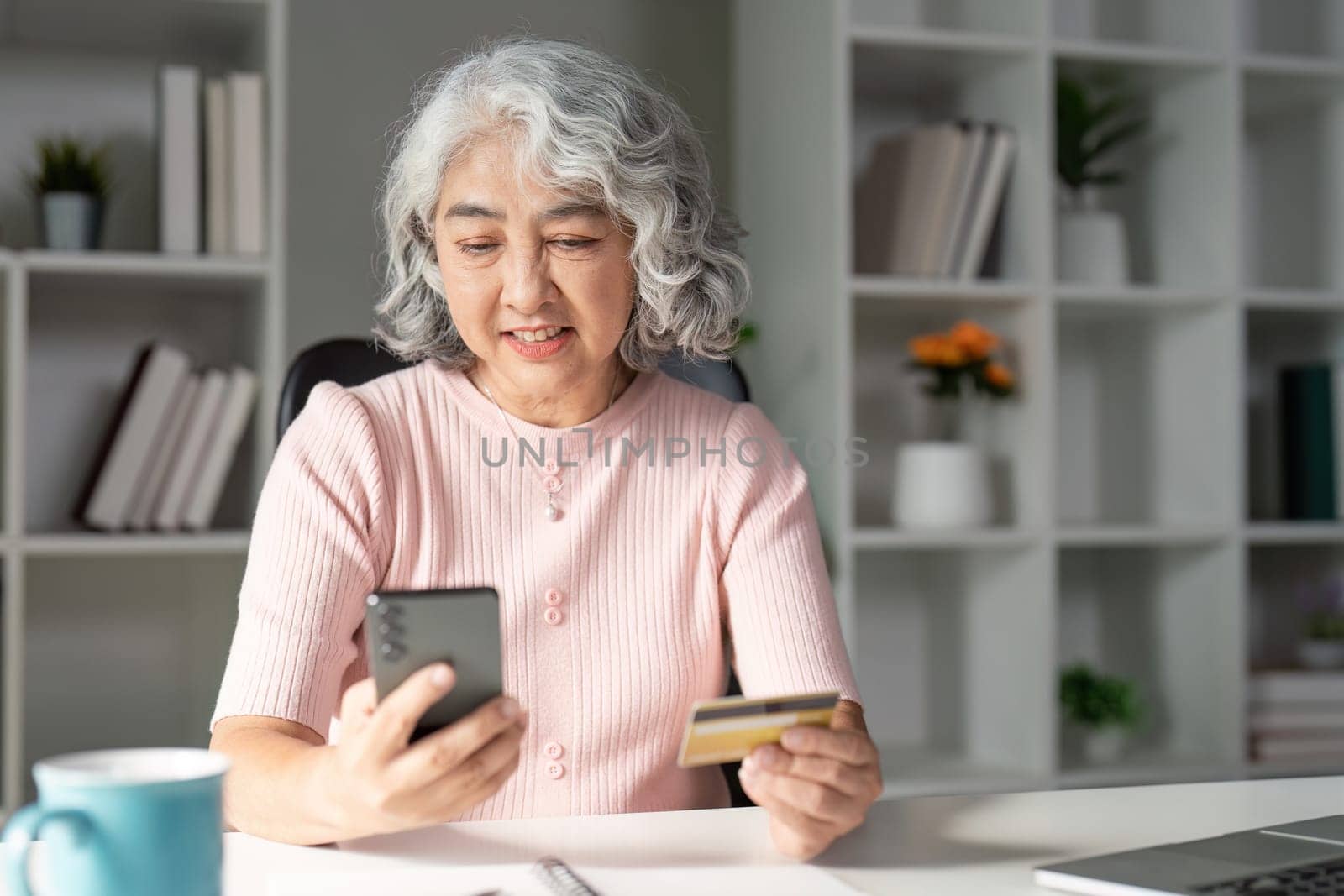 An elderly woman smiles while holding a credit card and using her smartphone for online shopping in a bright, inviting home office setting.